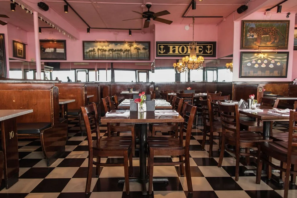 Empty restaurant with wooden tables and chairs, pink walls, chandeliers, black and white checkered floor, and various framed paintings.
