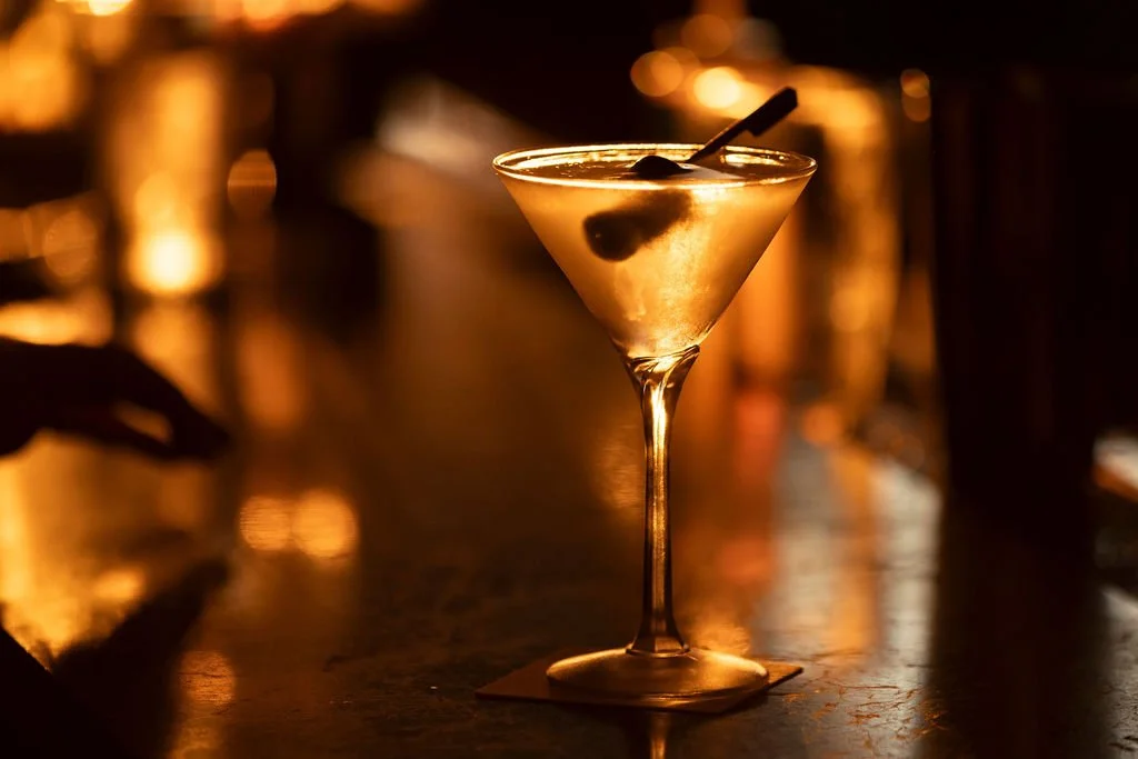 A cocktail glass with a lemon twist and cherry garnish on a bar counter, illuminated by warm, dim lighting.