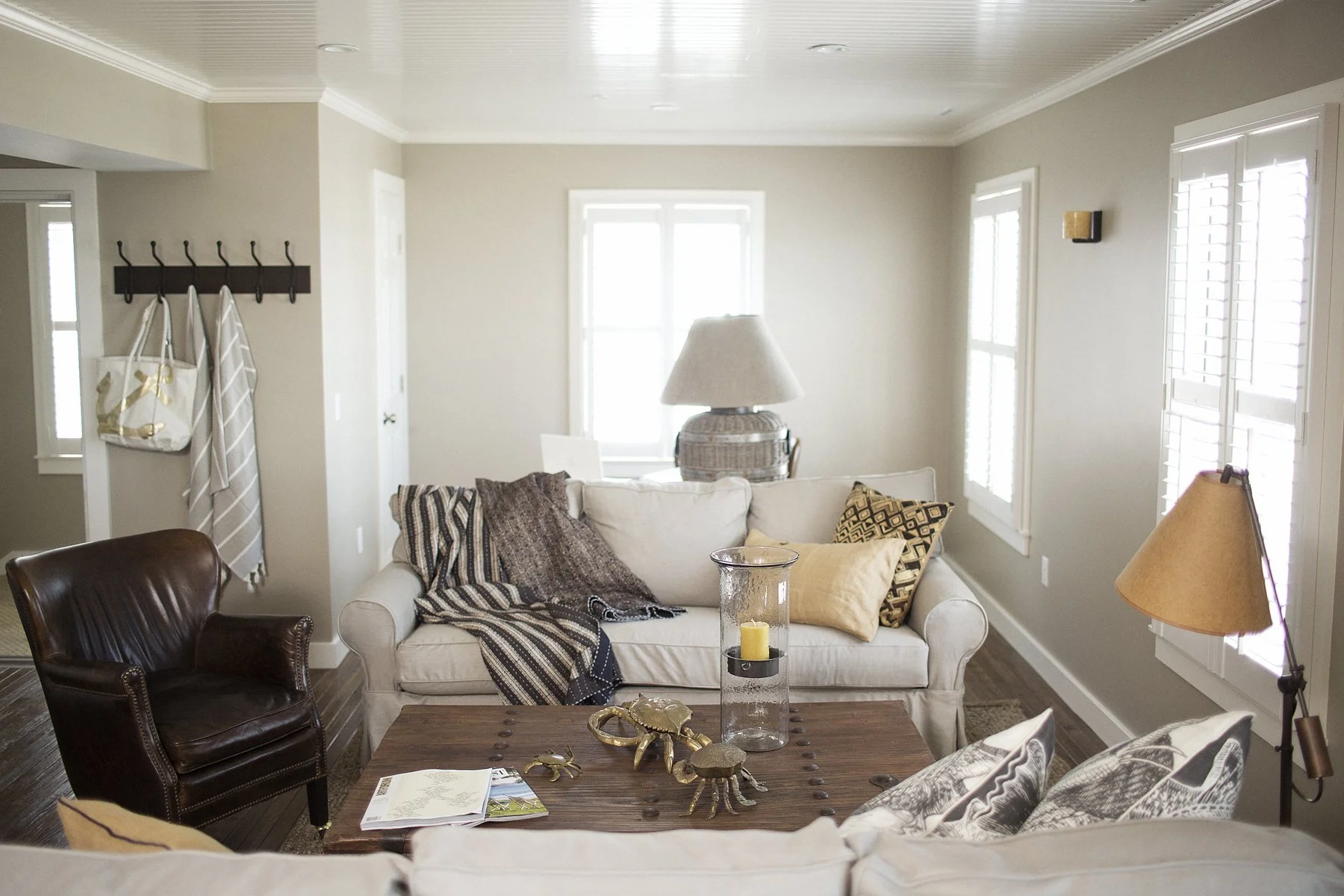 Living room with beige walls, white crown molding, a window with shutters, a white sofa with pillows and throws, a dark wooden coffee table with decorative items, a brown leather armchair, a side table with a lamp, and coat hooks with a bag and a str