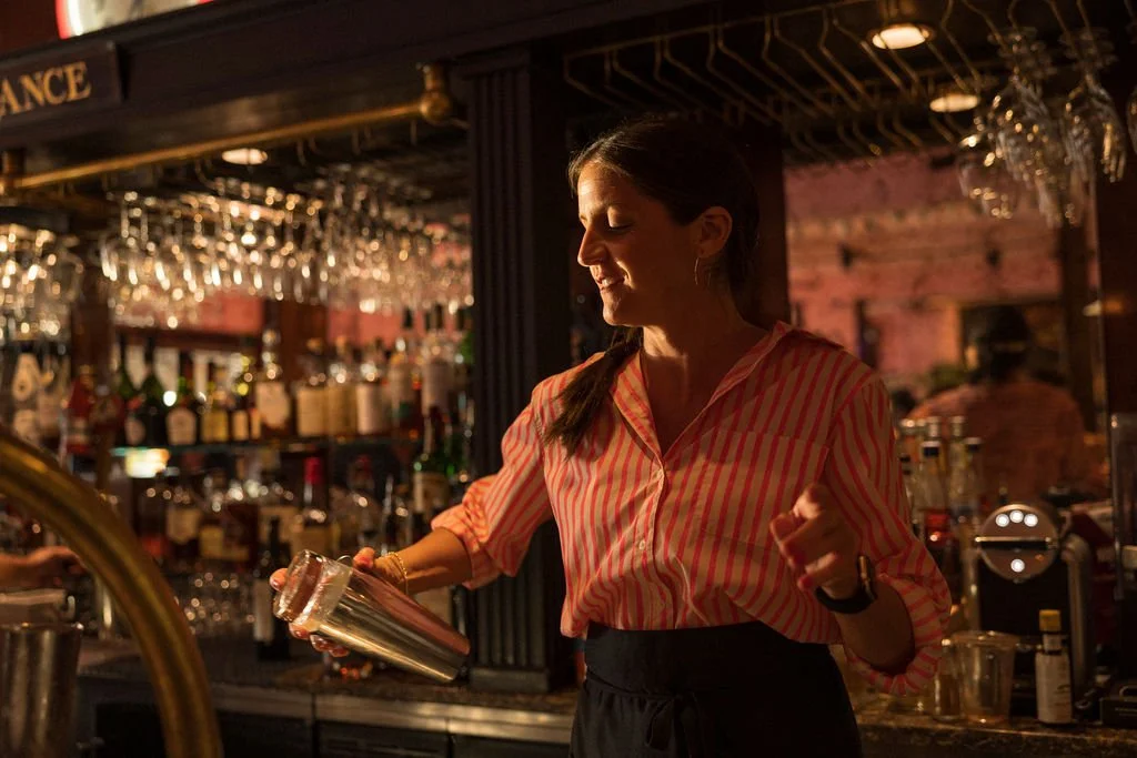 A woman behind a bar counter in a dimly lit bar, pouring a drink from a cocktail shaker, with shelves of liquor bottles and glasses in the background.