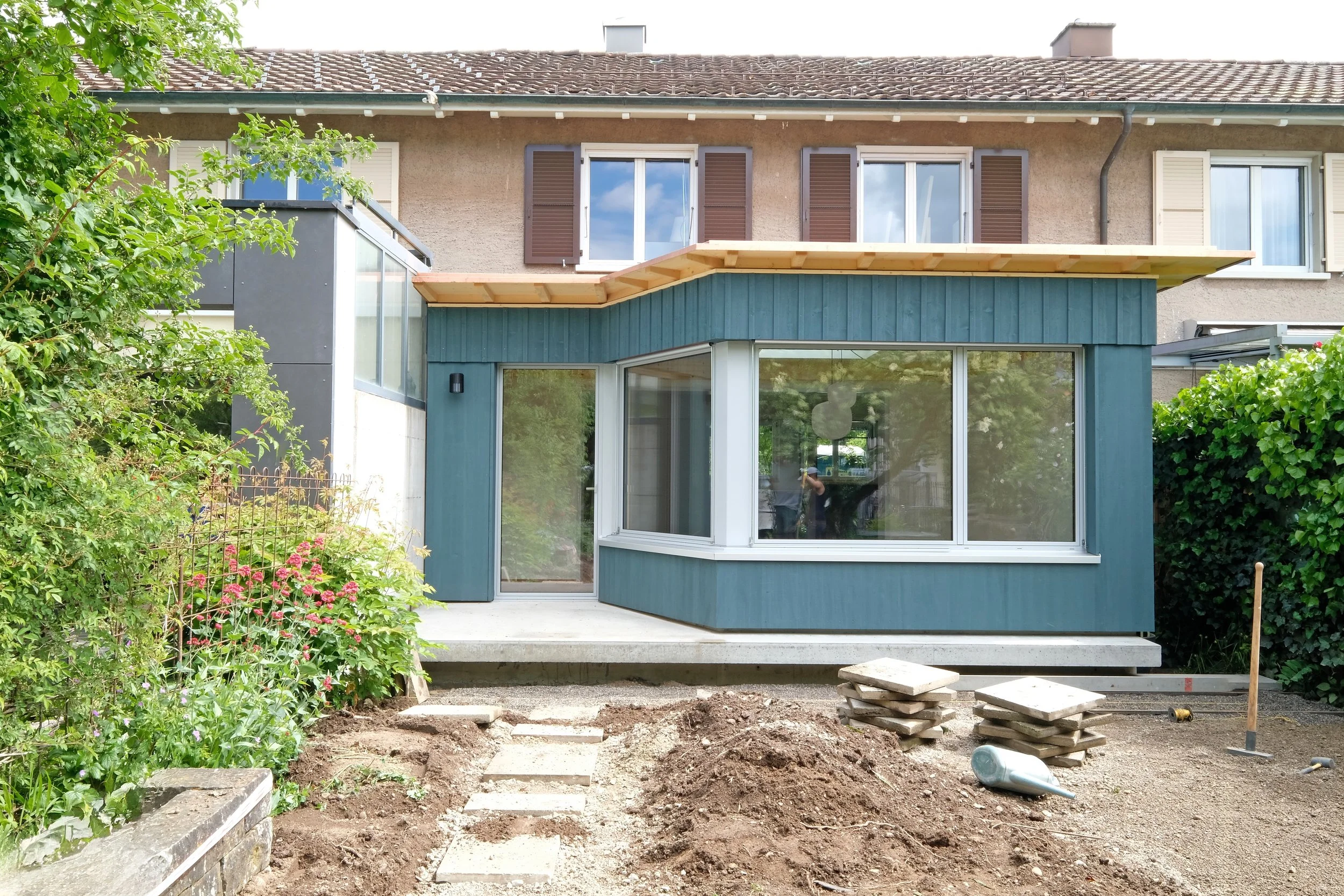 Ein zweistöckiges Haus mit einer neuen Terrasse im Bau, umgeben von Gartenpflanzen, im Vordergrund befinden sich Baustoffe und Erde.