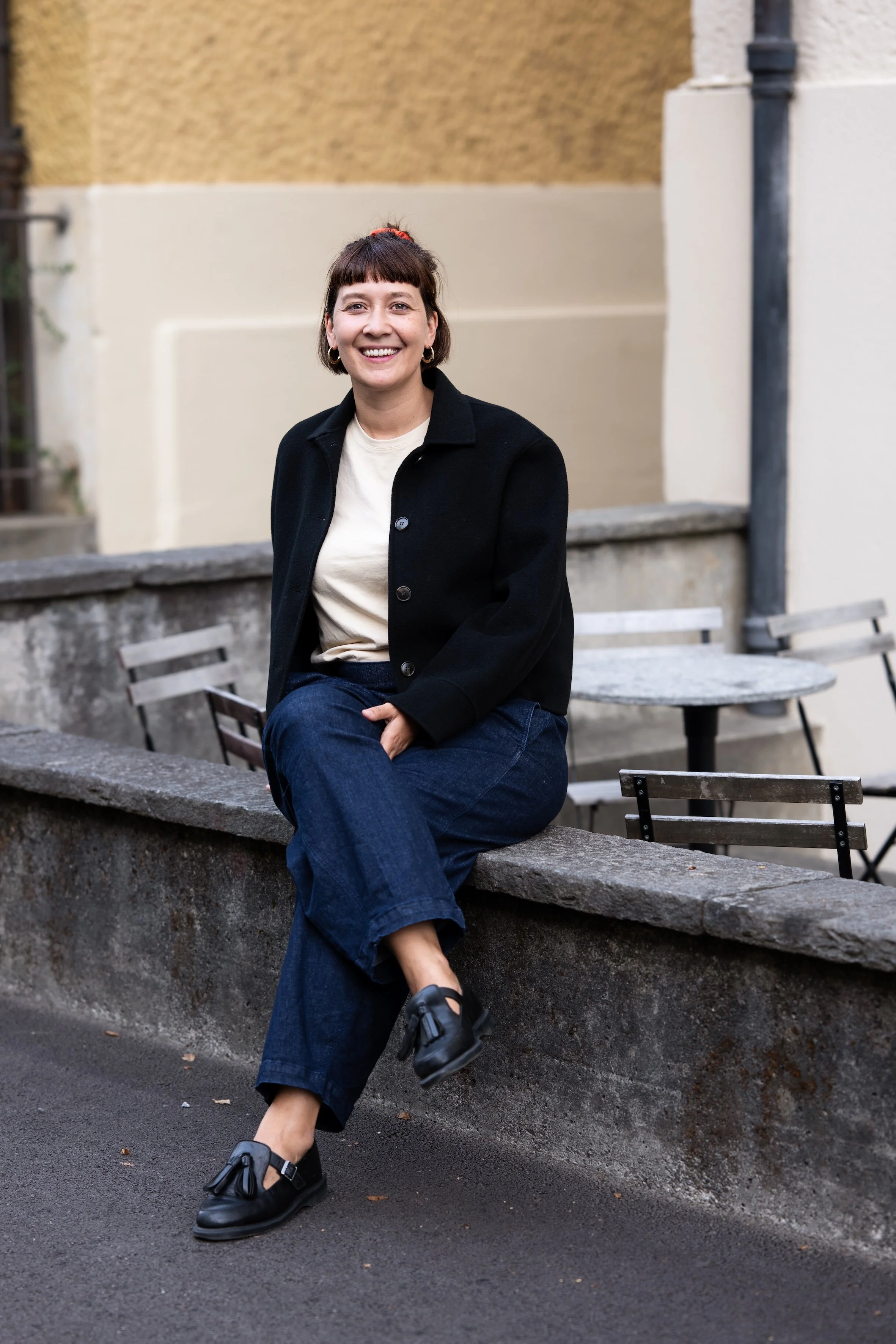 Eine lachende Frau mit kurzen dunklen Haaren, trägt eine schwarze Jacke, weißen T-Shirt, blaue Jeans, sitzt auf einer Mauer im Freien vor einer bunten Wand und einem Tisch.