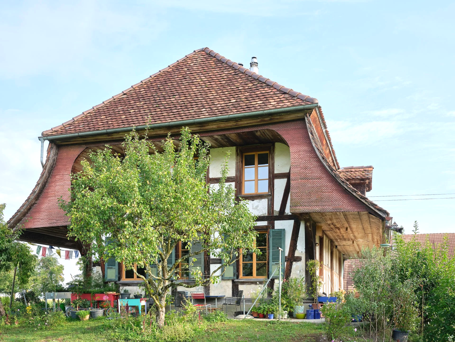 Ein idyllisches Haus mit traditioneller Architektur und einem begrünten Garten, voller Pflanzen und Gartenmöbel, unter blauem Himmel.