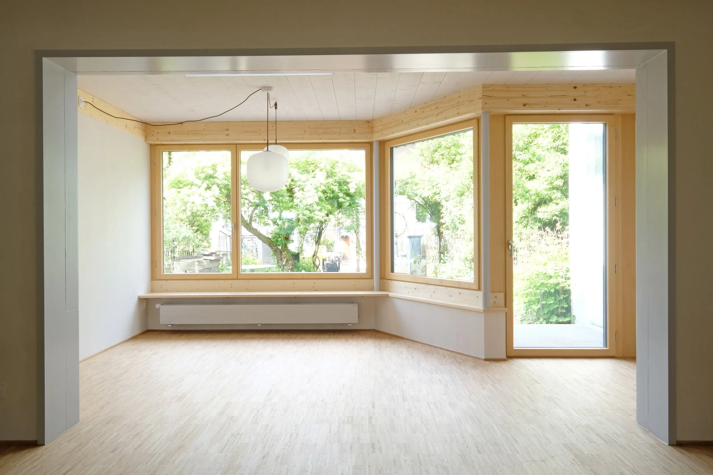 Innenansicht eines Zimmers mit großen Fenstern, Holzbalken und viel Tageslicht.