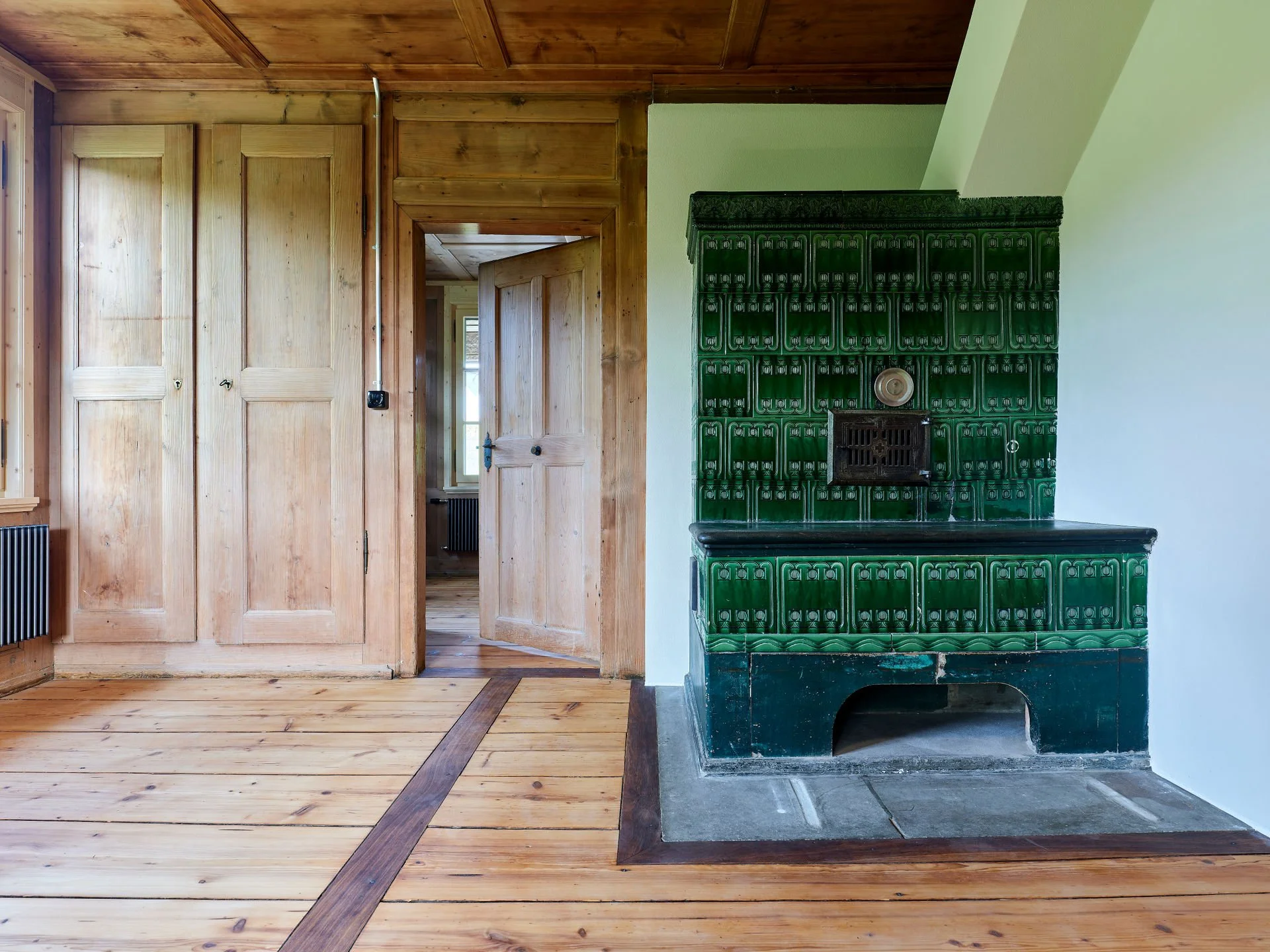 Ein Raum mit Holzboden, Holzrahmen und Türen, eine grüne Ofenfliese mit schwarzem Ofen, an der Wand auf der rechten Seite.