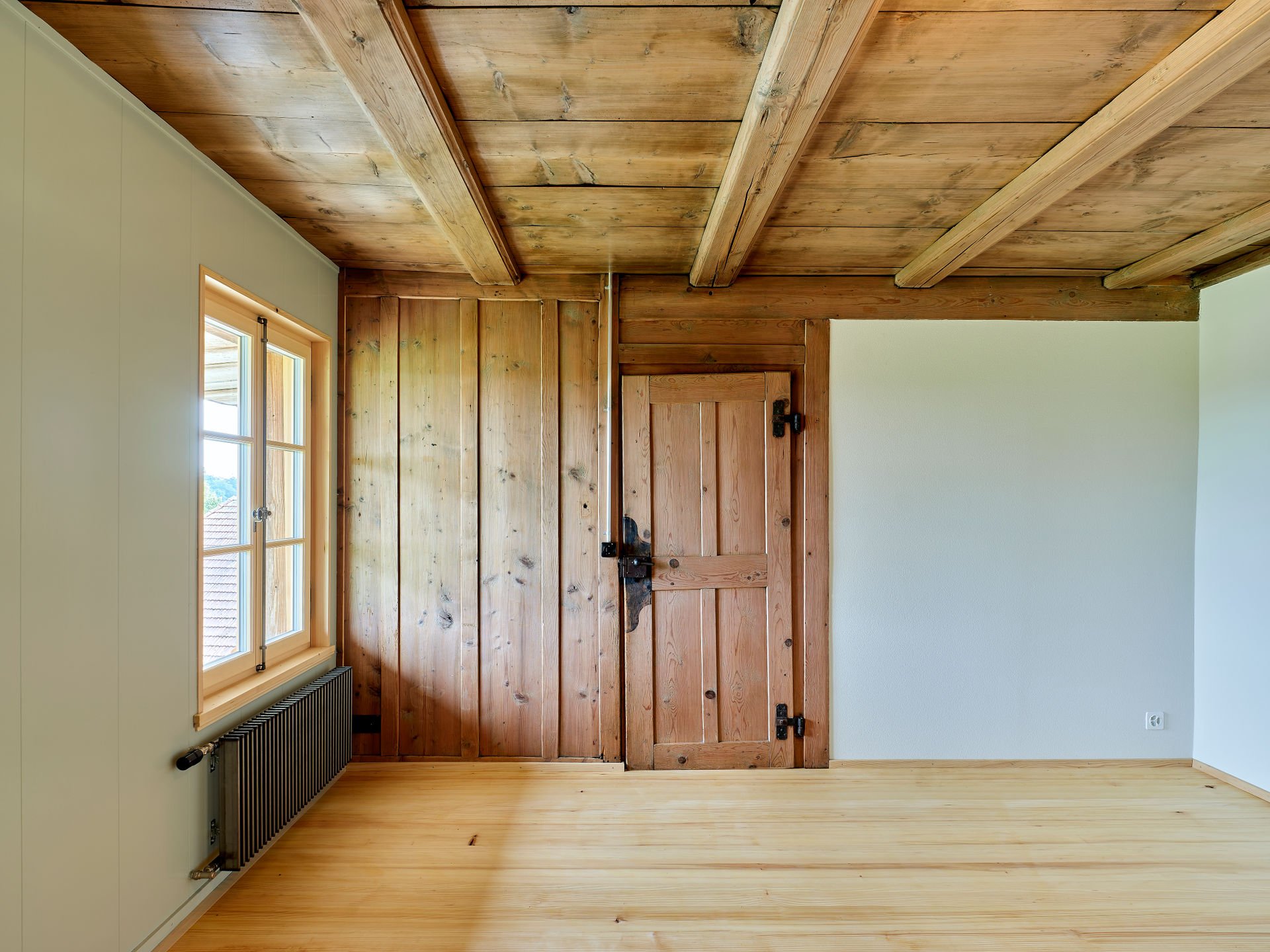 Innenansicht eines Raums mit Holzdecke, Holz Tür, einer weißen Wand und einem Fenster.