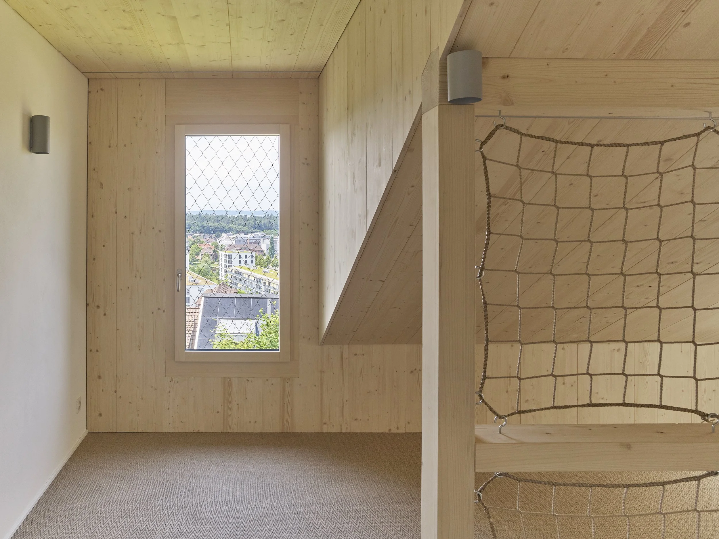Ein Zimmer mit Holzwänden, einem Fenster mit Blick auf eine Stadt, einem Kletterseil, einer Holzplattform, einem Netz und einer beige Teppichfläche.