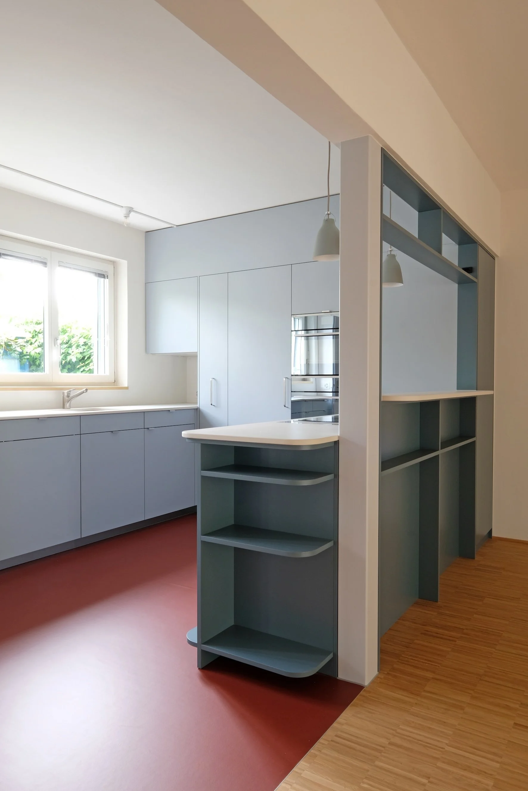 Innenansicht einer modernen Küche mit weißen Schränken, einer Kücheninsel und offenen Regalen an einer Wand, große Fenster lassen viel natürliches Licht herein.