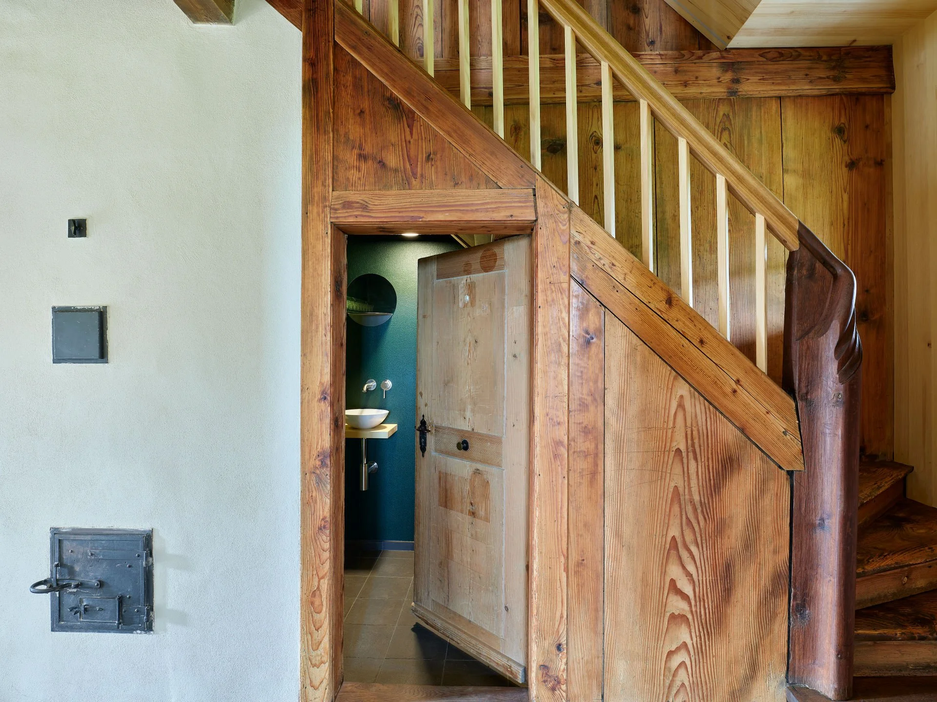 Innenansicht eines Hauses mit Holztreppen, einer Tür zu einer Toilette, einem Waschbecken und einem Spiegel.