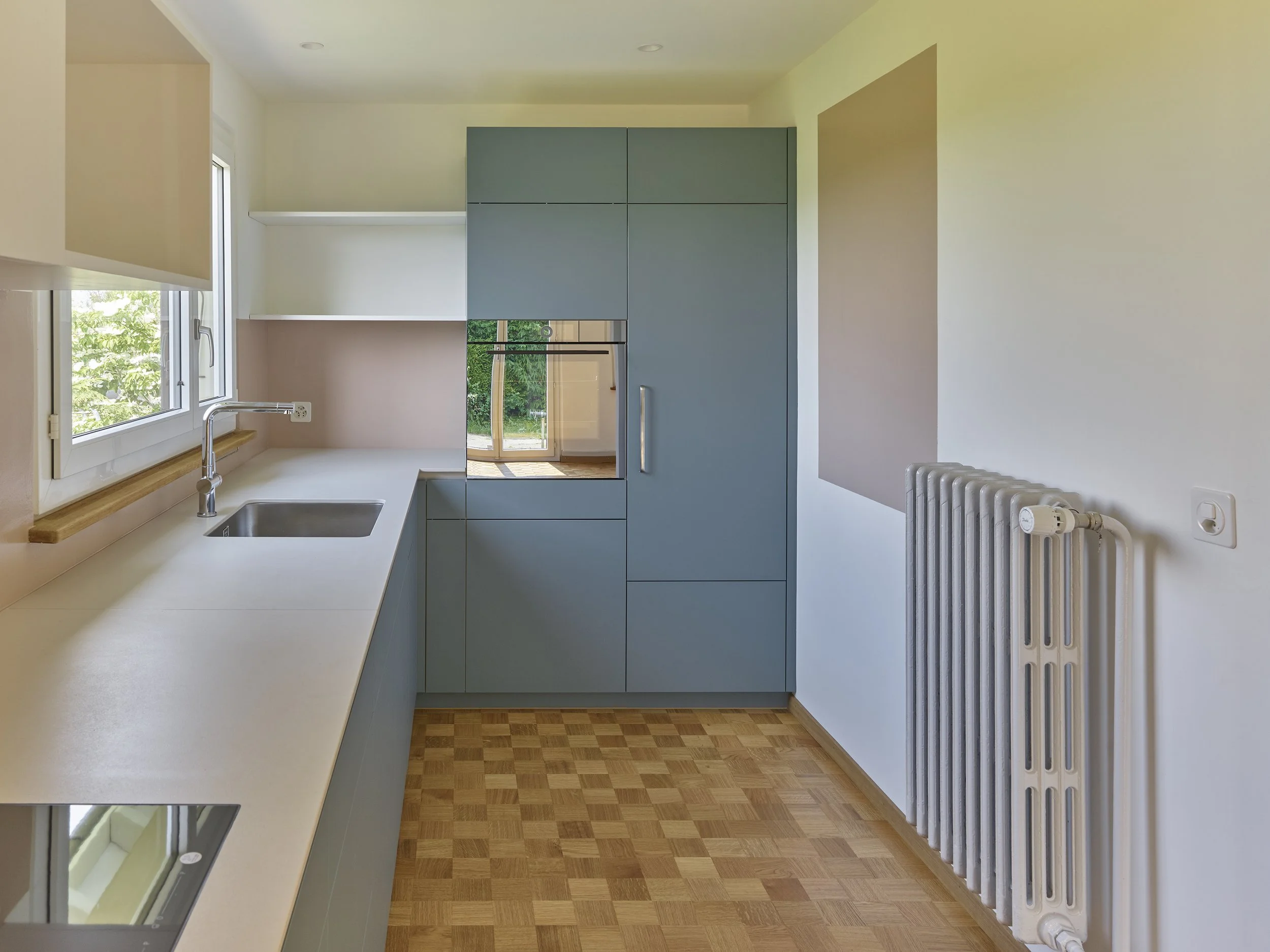 Moderne Küche mit weißer Arbeitsplatte, blauen Schränken, Fenster über der Spüle und einem Heizkörper an der Wand.