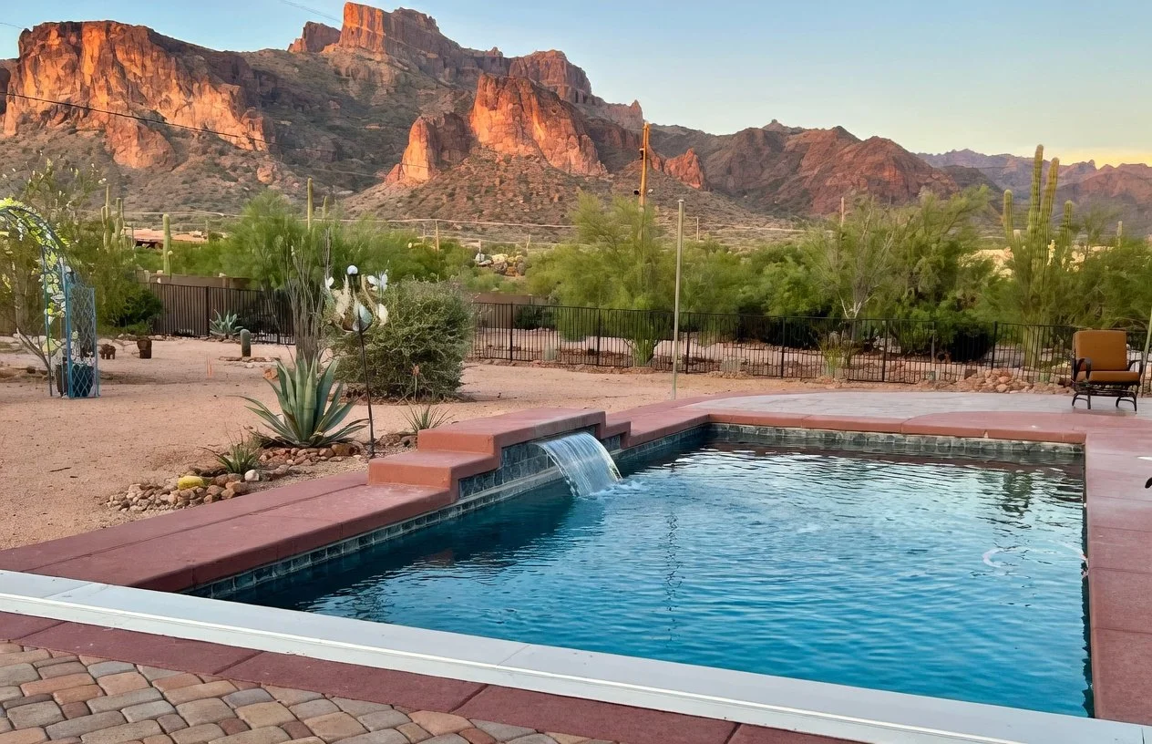 Swimming pool with a small waterfall feature, surrounded by a paved patio and desert landscaping, with cacti and desert plants in the yard, and mountains in the background at sunset.