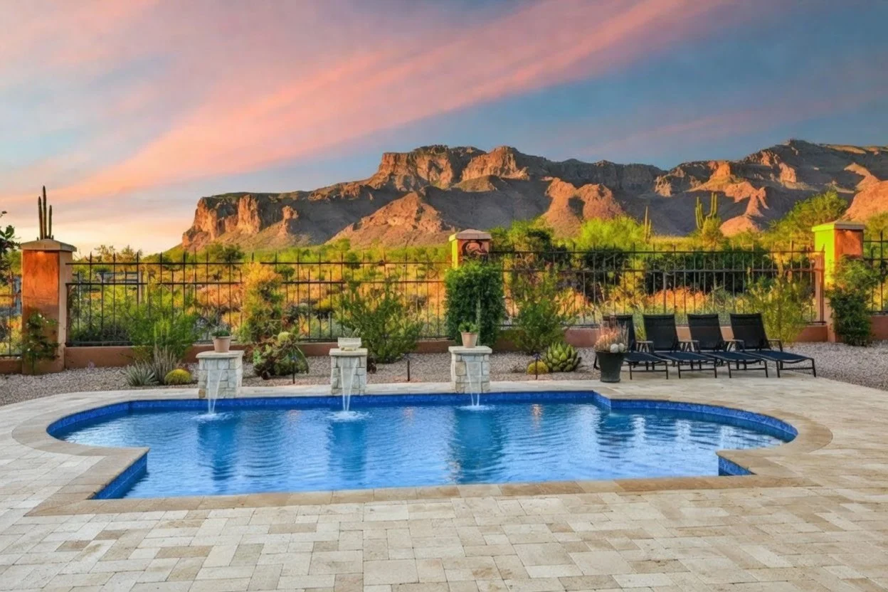 A swimming pool with water features, surrounded by a patio with lounge chairs, set against a desert landscape with mountains and cacti at sunset.