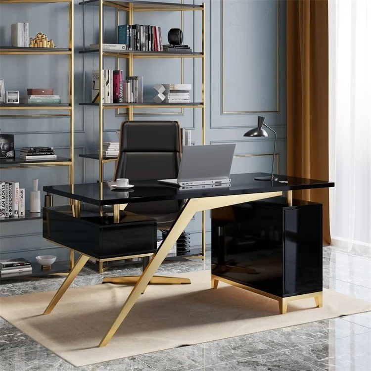 Modern office desk with a black surface, gold angled legs, and black storage units on each side. Behind the desk is a black office chair. To the right of the desk is a sleek black desk lamp. The background features a blue wall with gold trim and large gold shelving filled with books and decorative items. A window with white sheer curtains and gold drapes is on the far right.