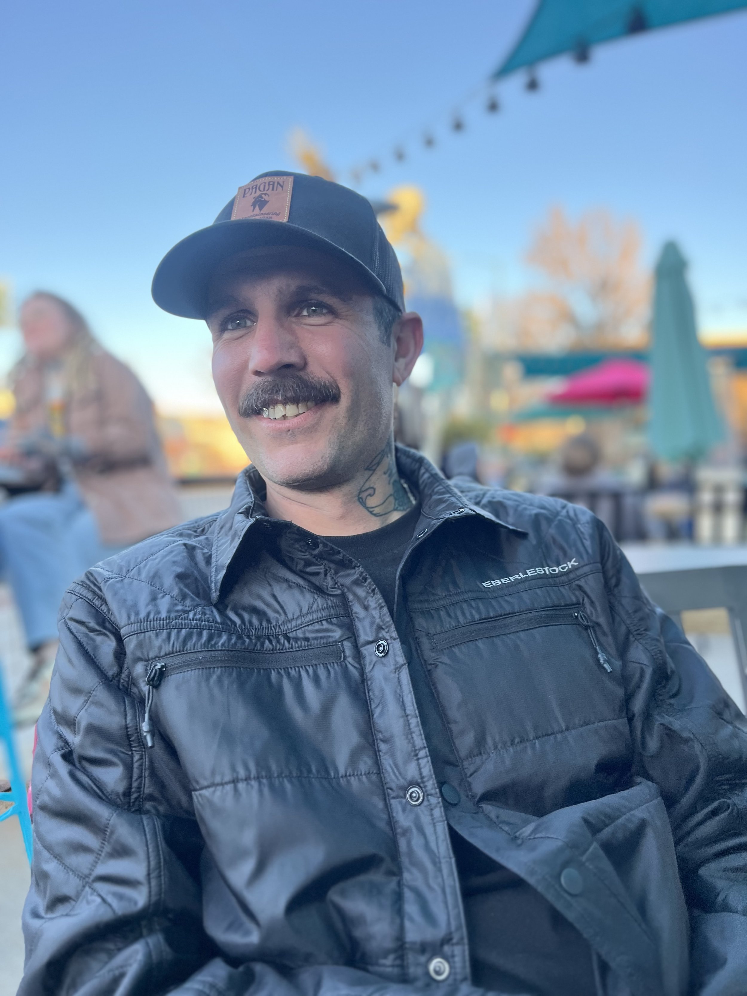 A man with a mustache and tattoos is smiling, wearing a dark jacket and a baseball cap, sitting outdoors with blue sky, a person in the background, and umbrellas with string lights.
