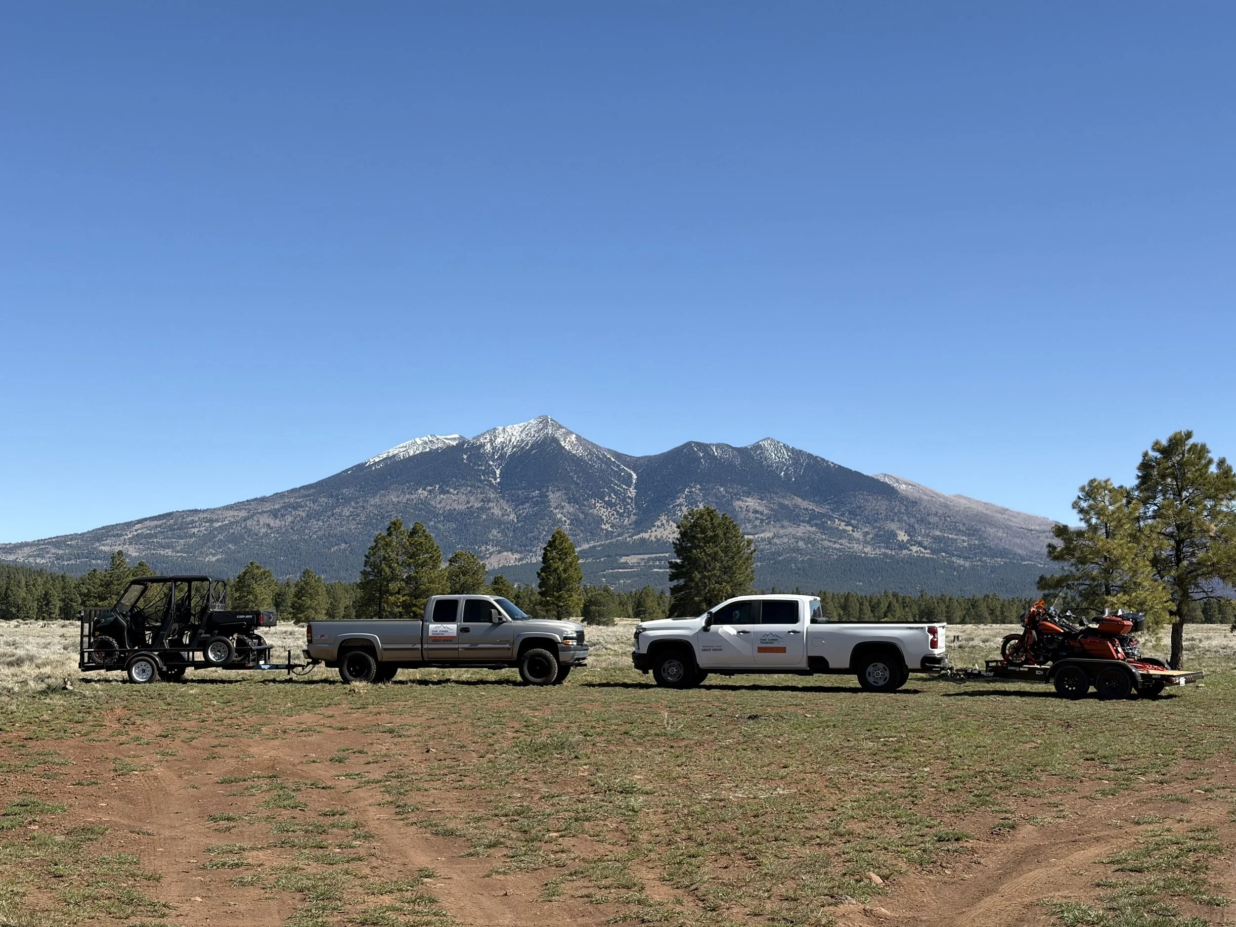 Vehicles including a utility truck, a trailer, and three motorcycles parked on a grassy field with mountains in the background and a clear blue sky.