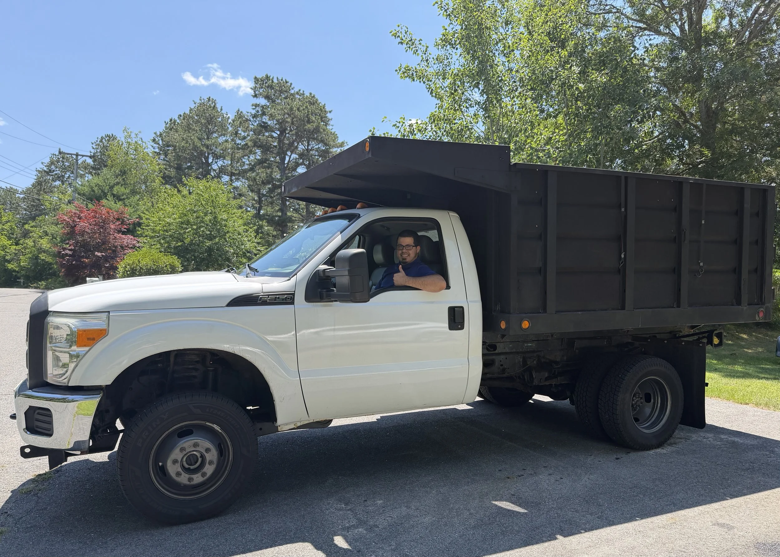 A man smiling and giving a thumbs-up while sitting inside a white Ford F-350 pickup truck with a black cargo area, parked outside surrounded by trees and blue sky.