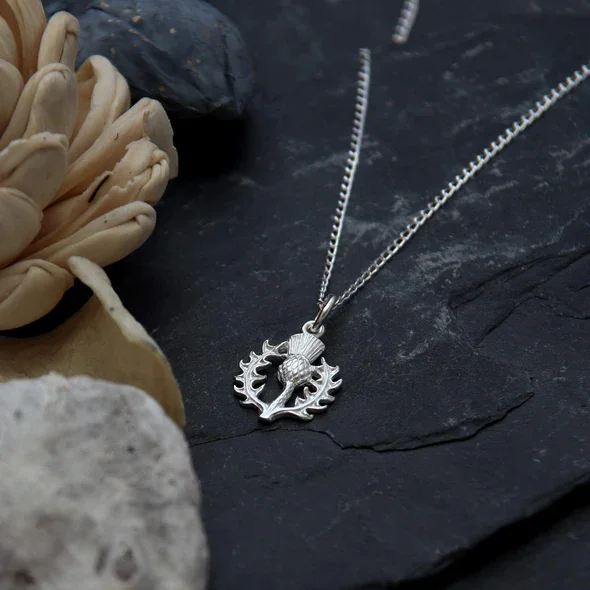 Sterling Silver Scottish Thistle Pendant, feathered head, crosshatched globe, open spiky leaf surround, shown on Scottish slate with sea stones and dried botanicals