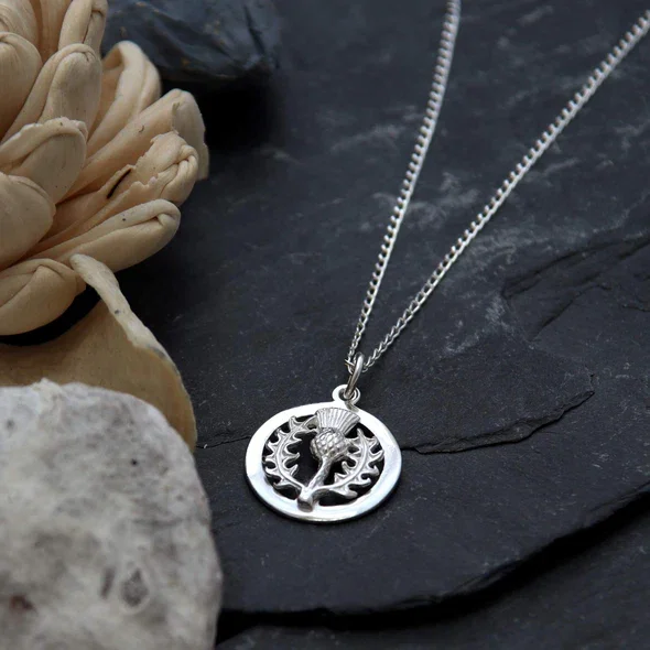 Sterling Silver Scottish Thistle Pendant, circular frame, feathered head, crosshatched globe, spiky leaves, shown on Scottish slate with sea stones and dried botanicals