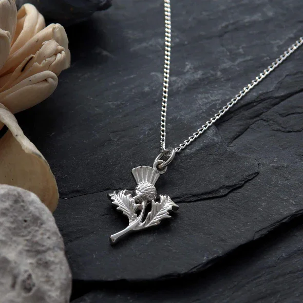Sterling Silver Scottish Thistle Pendant, feathered head, crosshatched globe, naturalistic leaf detail, shown on Scottish slate with sea stones and dried botanicals