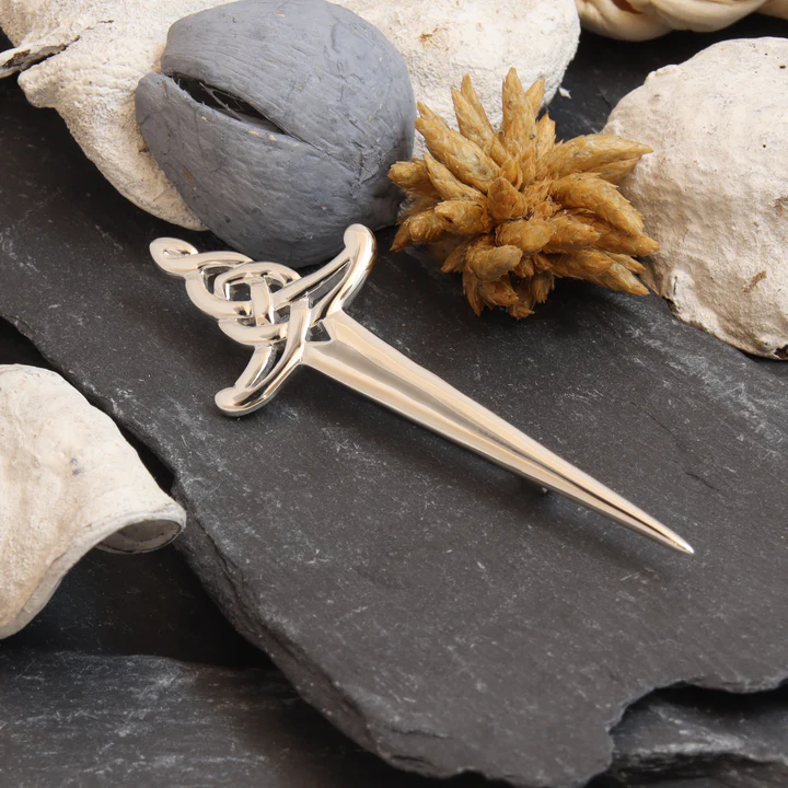 Sterling Silver Scottish sword kilt pin with Celtic knotwork hilt, displayed on dark Scottish slate with natural sea stones and dried botanicals