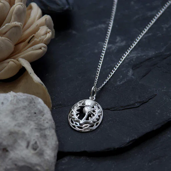 Sterling Silver Scottish Thistle Pendant, open oval frame, feathered head, crosshatched globe, pierced leaf detail, shown on Scottish slate with sea stones and dried botanicals