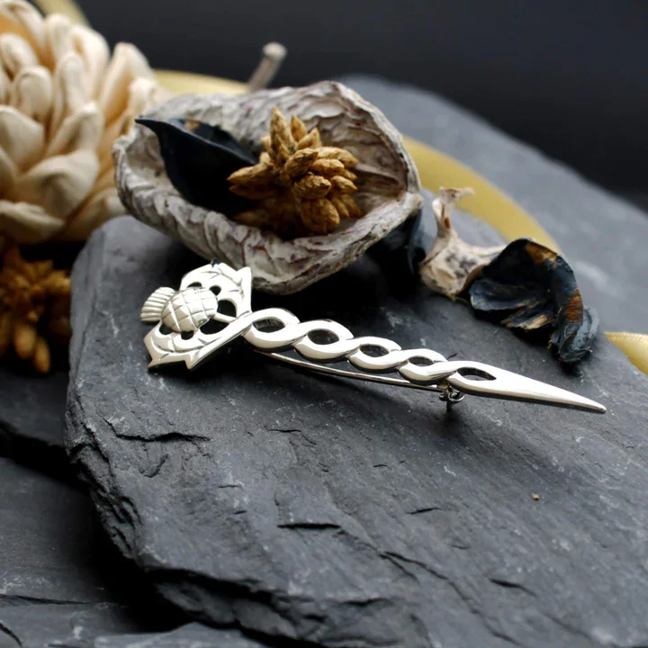 Sterling Silver Scottish Thistle Kilt Pin with Celtic knotwork stem, displayed on dark Scottish slate with dried botanicals