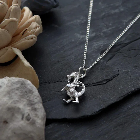 Sterling Silver Nessie Bagpiper Pendant, Loch Ness Monster playing bagpipes, sculpted novelty form, shown on Scottish slate with sea stones and dried botanicals