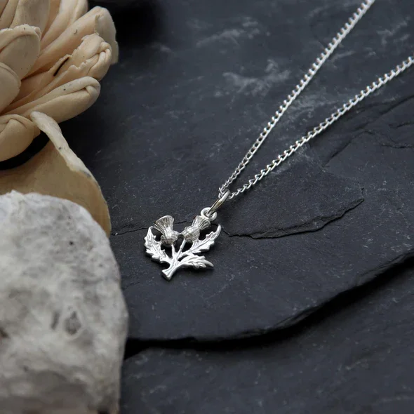 Sterling Silver Scottish Thistle Pendant, twin-head botanical form, feathered crowns, crosshatched globes, shown on Scottish slate with sea stone and dried botanicals