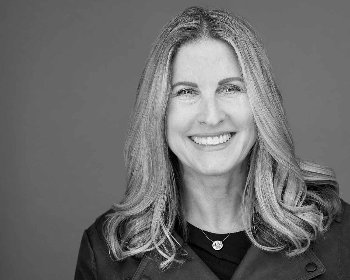 Black and white portrait of a smiling woman with long wavy hair, wearing a dark jacket and a necklace, against a plain background.