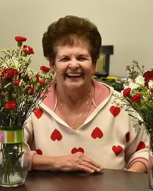 A smiling woman sitting at a table with red and white flowers in vases on either side of her, wearing a white sweater with red hearts.