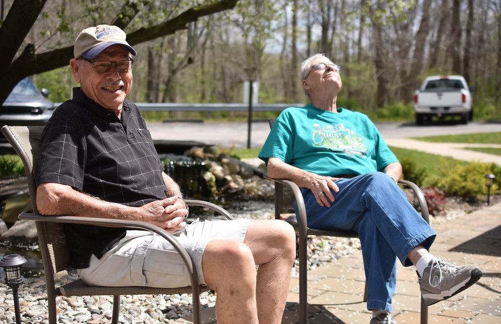 An elderly man and woman sitting outdoors on patio chairs under a tree, smiling and relaxing in bright sunlight.