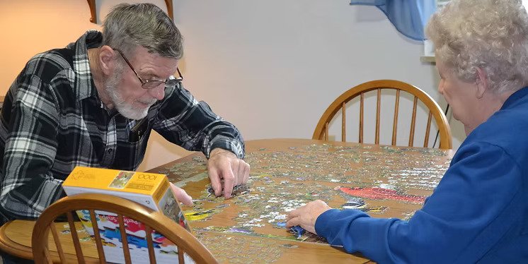 An elderly man and woman sitting at a wooden table, working on assembling a jigsaw puzzle together.