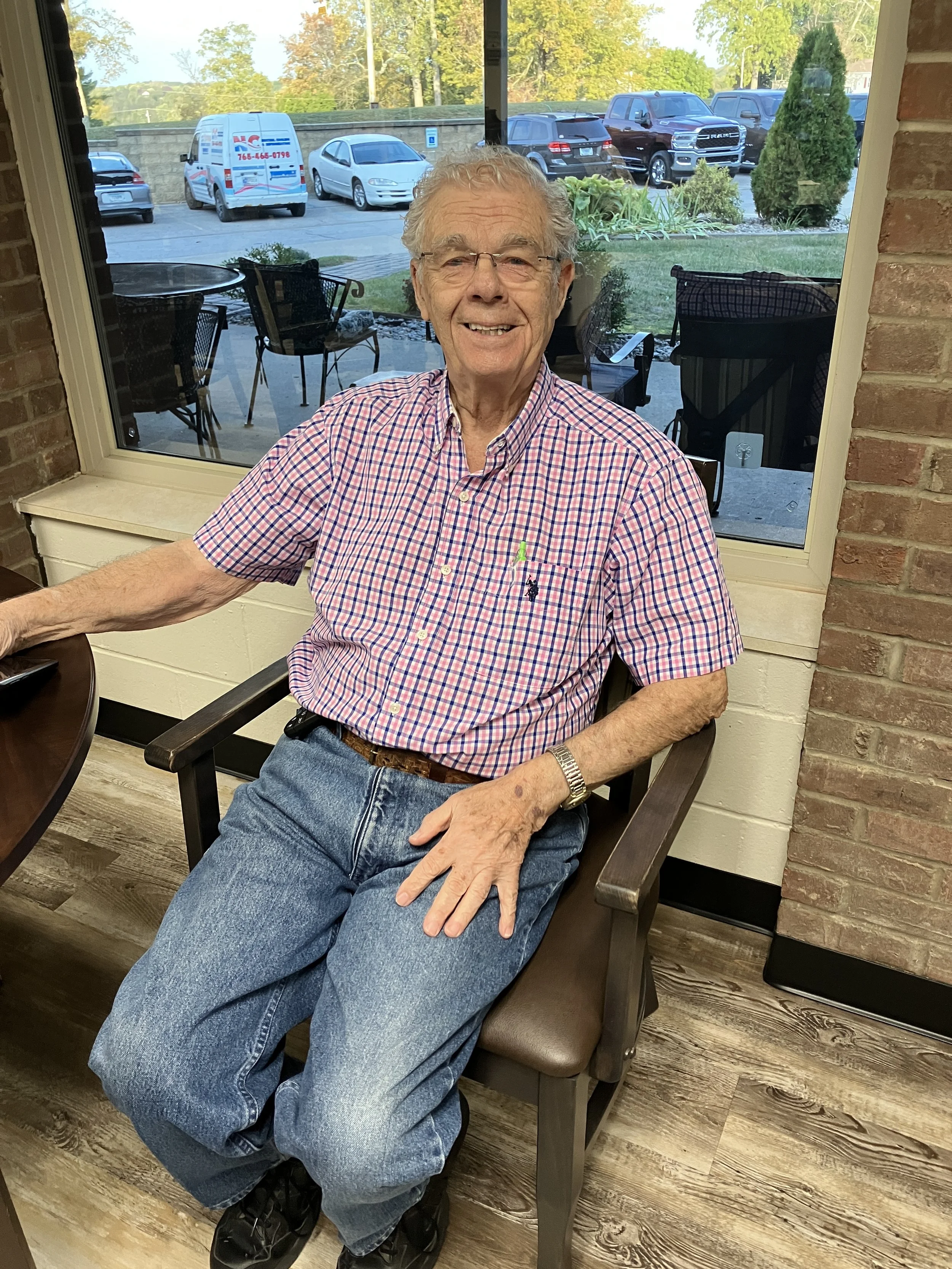 An elderly man with glasses smiling and sitting in a chair indoors near a large window, wearing a red and white checkered shirt, blue jeans, and a wristwatch.