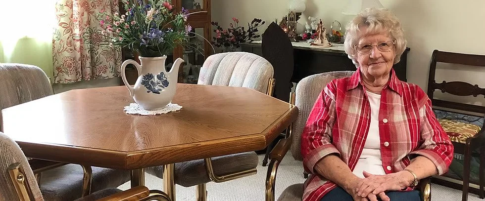 An elderly woman with curly white hair and glasses sitting at a dining table in a living room. She is wearing a red plaid shirt over a white top and is smiling. On the table, there is a large white ceramic pitcher with blue floral designs, filled with purple and green flowers. The background shows decorated shelves and a vintage-style curtain.