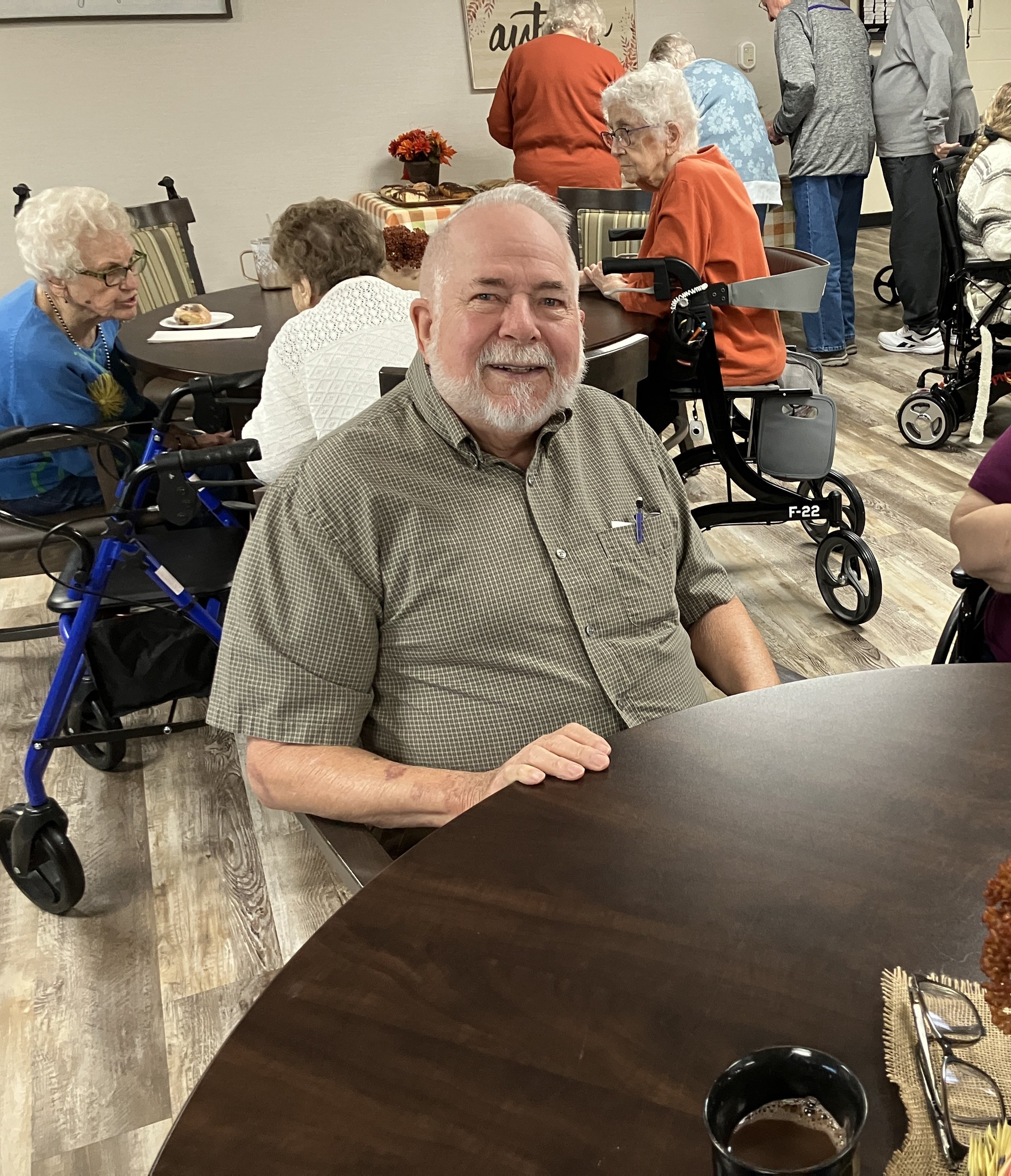 A smiling elderly man with a beard and light hair sitting at a table in a social gathering at a senior center or similar facility.