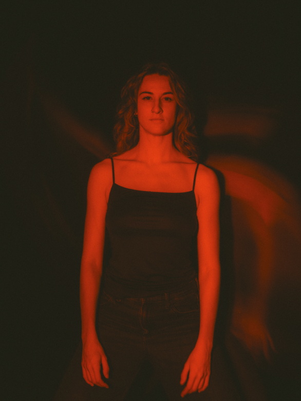 Une jeune femme avec des cheveux bouclés et une expression sérieuse, portant un haut noir à fines bretelles, se tient devant un fond sombre avec un éclairage rouge.