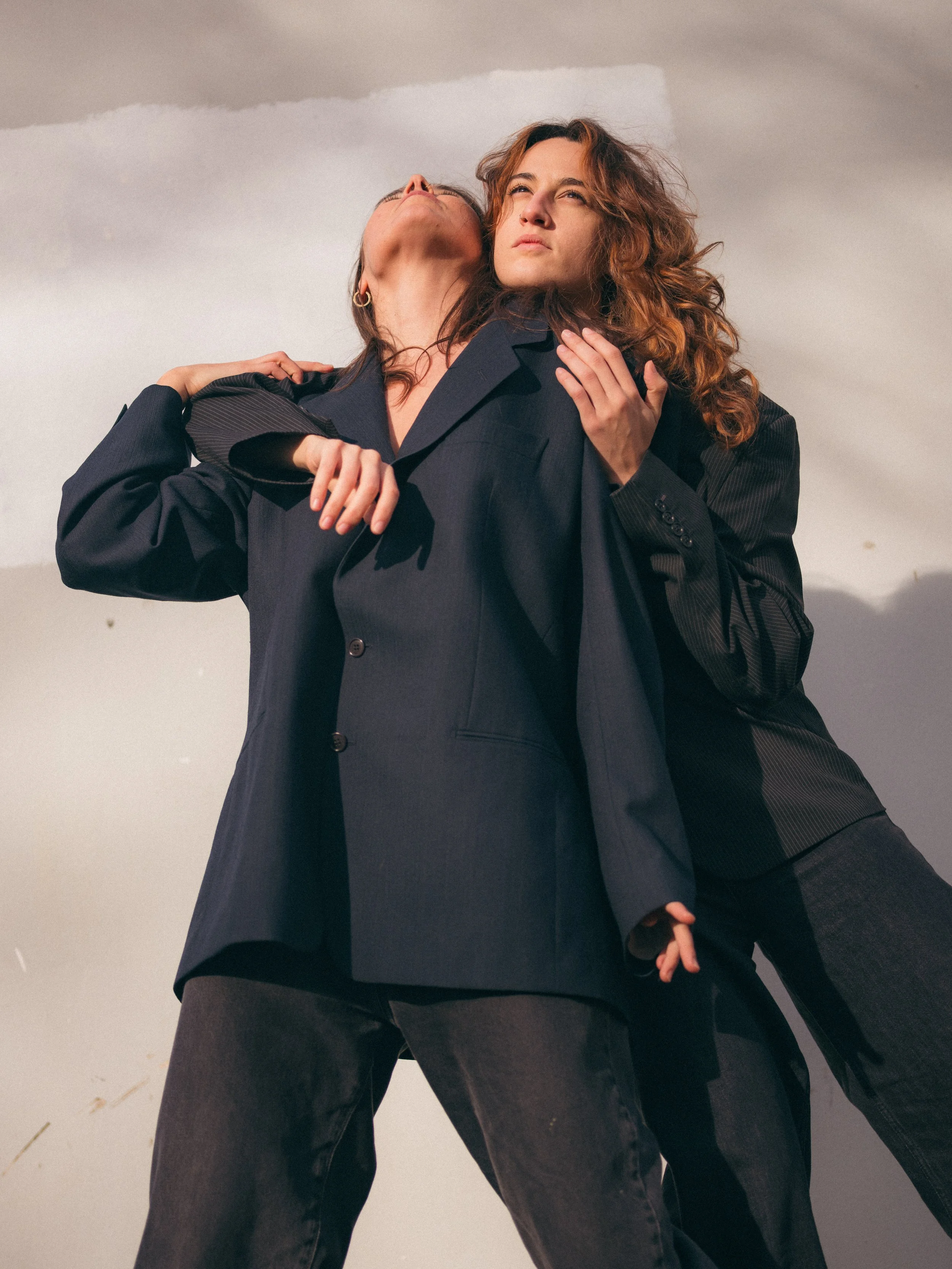 Deux femmes en costume noir debout côte à côte, l'une regarde vers le haut et l'autre regarde vers l'avant, dans un ciel nuageux.