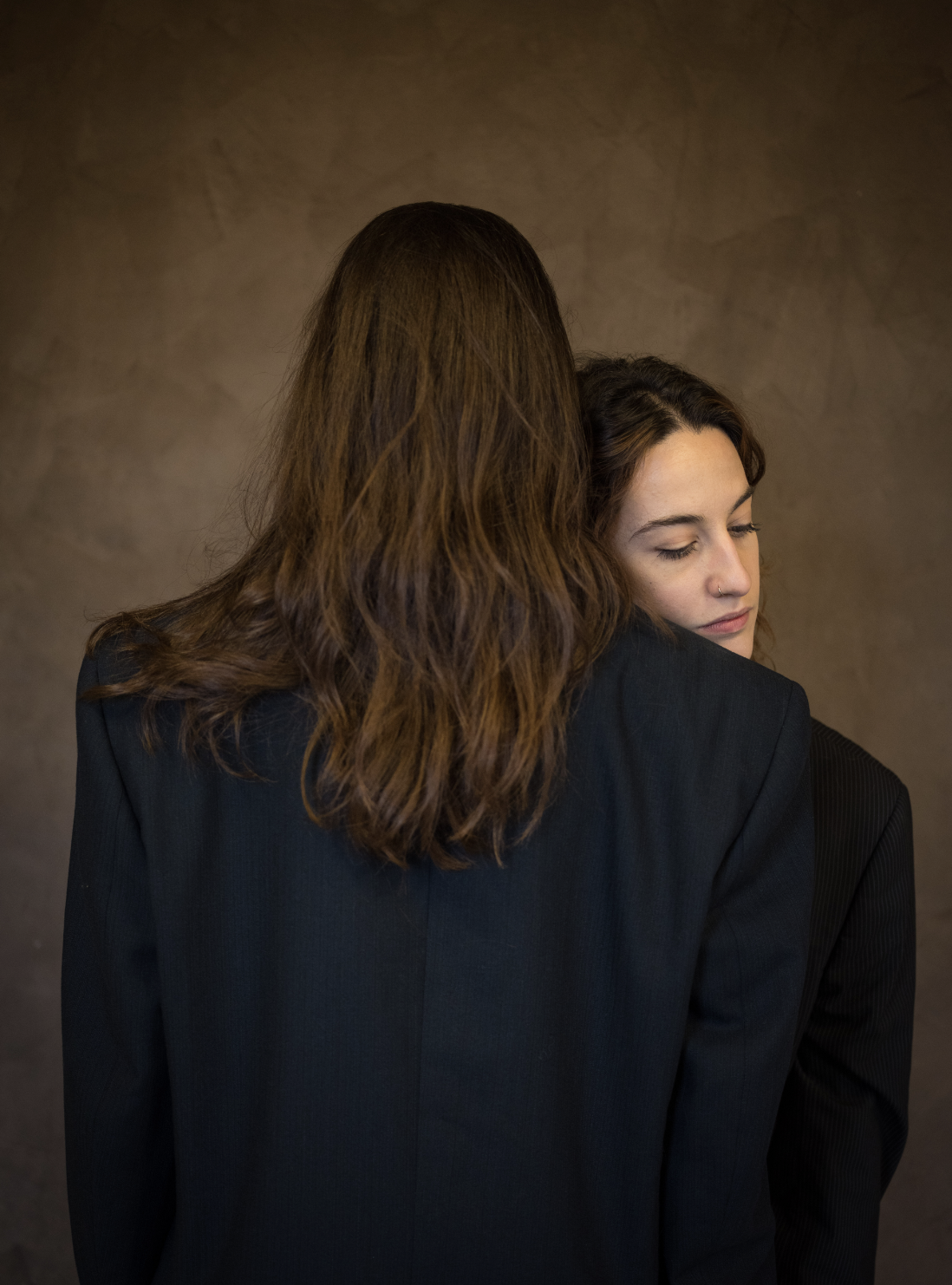 Deux femmes habillées en noir, l'une avec les cheveux longs et bruns, l'autre avec les cheveux courts et bouclés, en train de se faire un câlin contre un fond uni