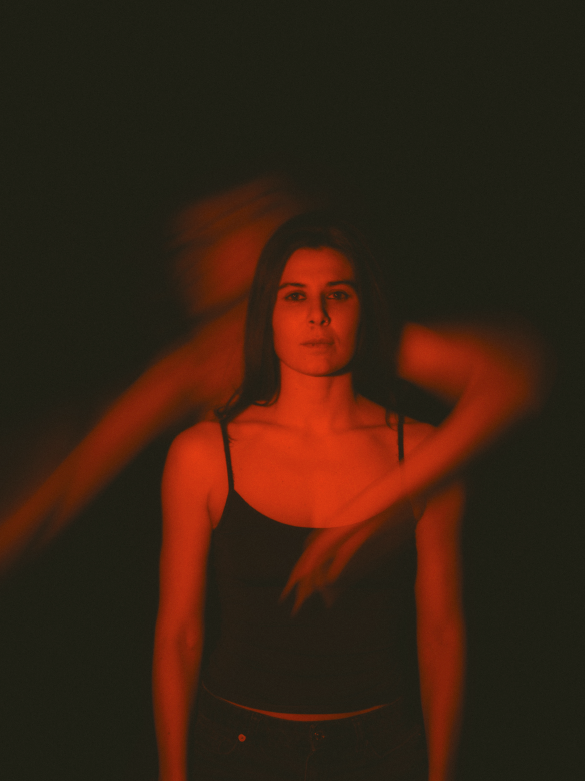 Jeune femme avec cheveux longs en haut noir, fond sombre, mouvement flou rouge orange autour, expression neutre, position droite, lumière faible