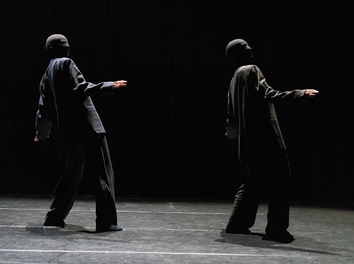 Deux personnes enveloppées de noir avec des masques noirs, marchant dans un espace sombre. Elles portent des vêtements décontractés et semblent être en mouvement.