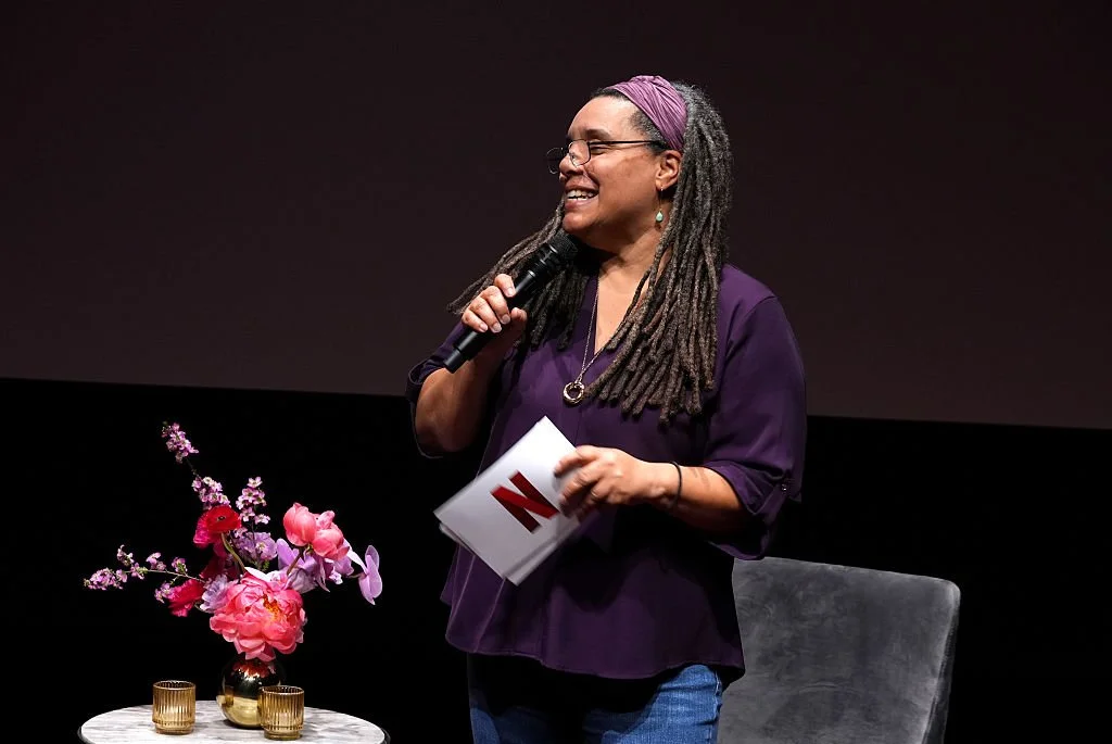 A woman with long dreadlocks, wearing glasses, a purple headband, a purple top, and jeans, is smiling and speaking into a microphone on stage. There is a table with pink flowers and candles beside her.