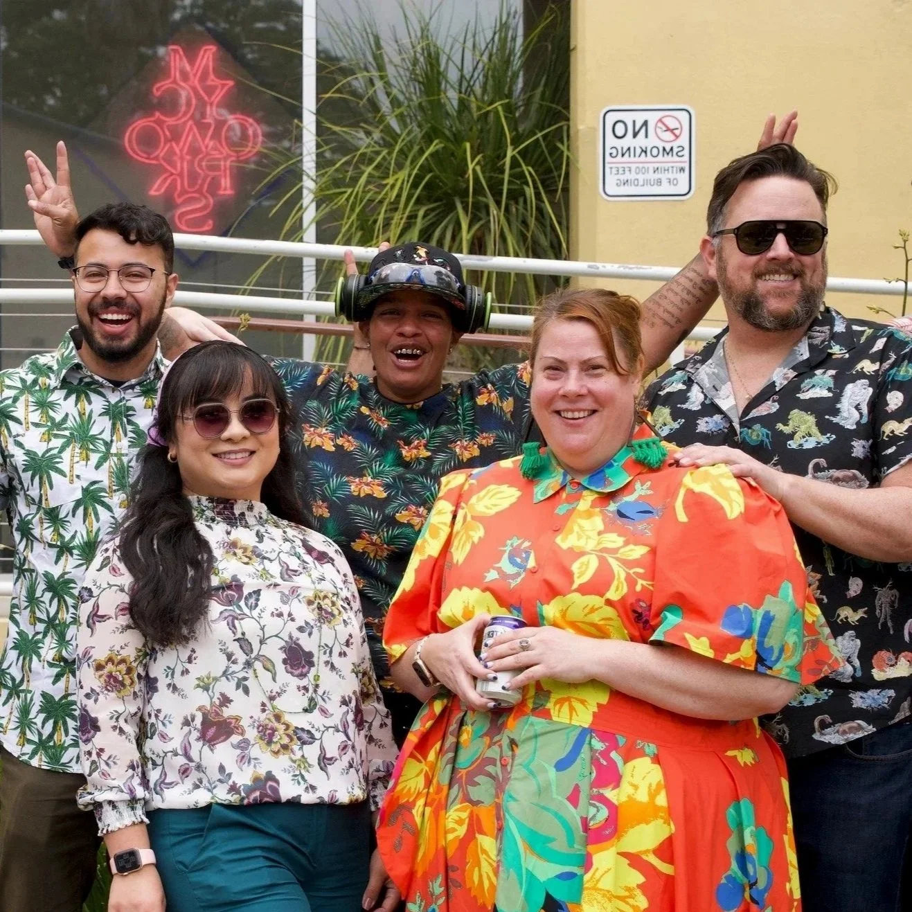 Five people standing outdoors wearing colorful, tropical-themed clothing, smiling and posing together.