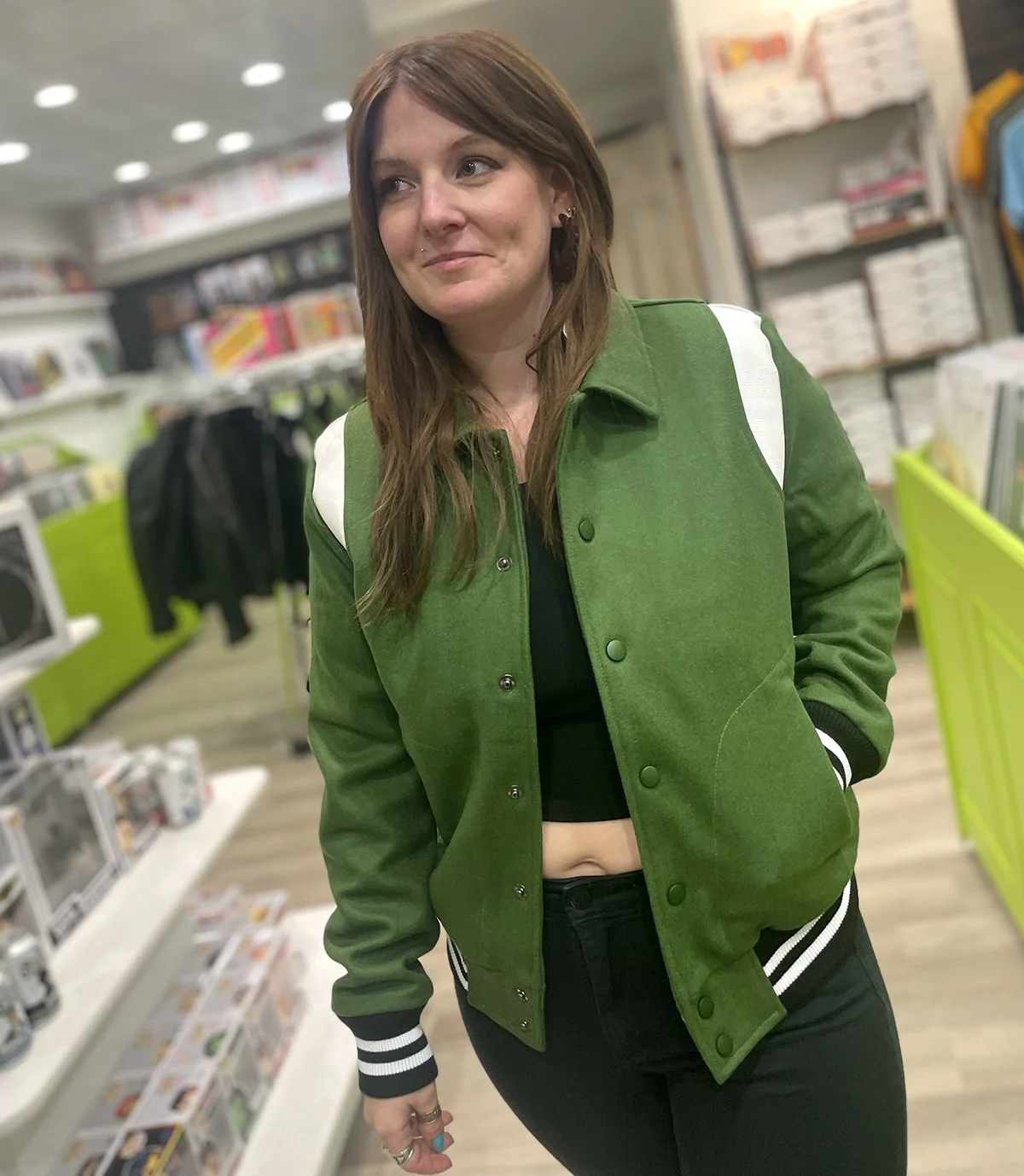 A woman with brown hair wearing a green varsity jacket with white accents, a black crop top, and black pants, standing in a store aisle with shelves of electronics and other items in the background.