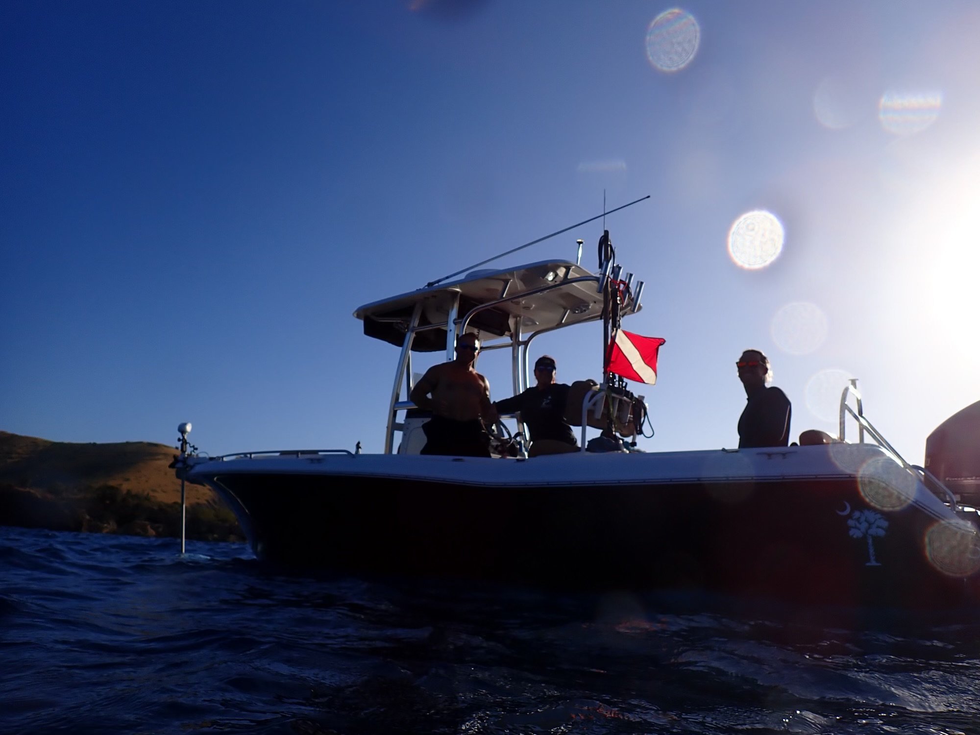 Silhouettes of three people on a boat at sunset, with hilly shoreline in the background and glare from the sun.