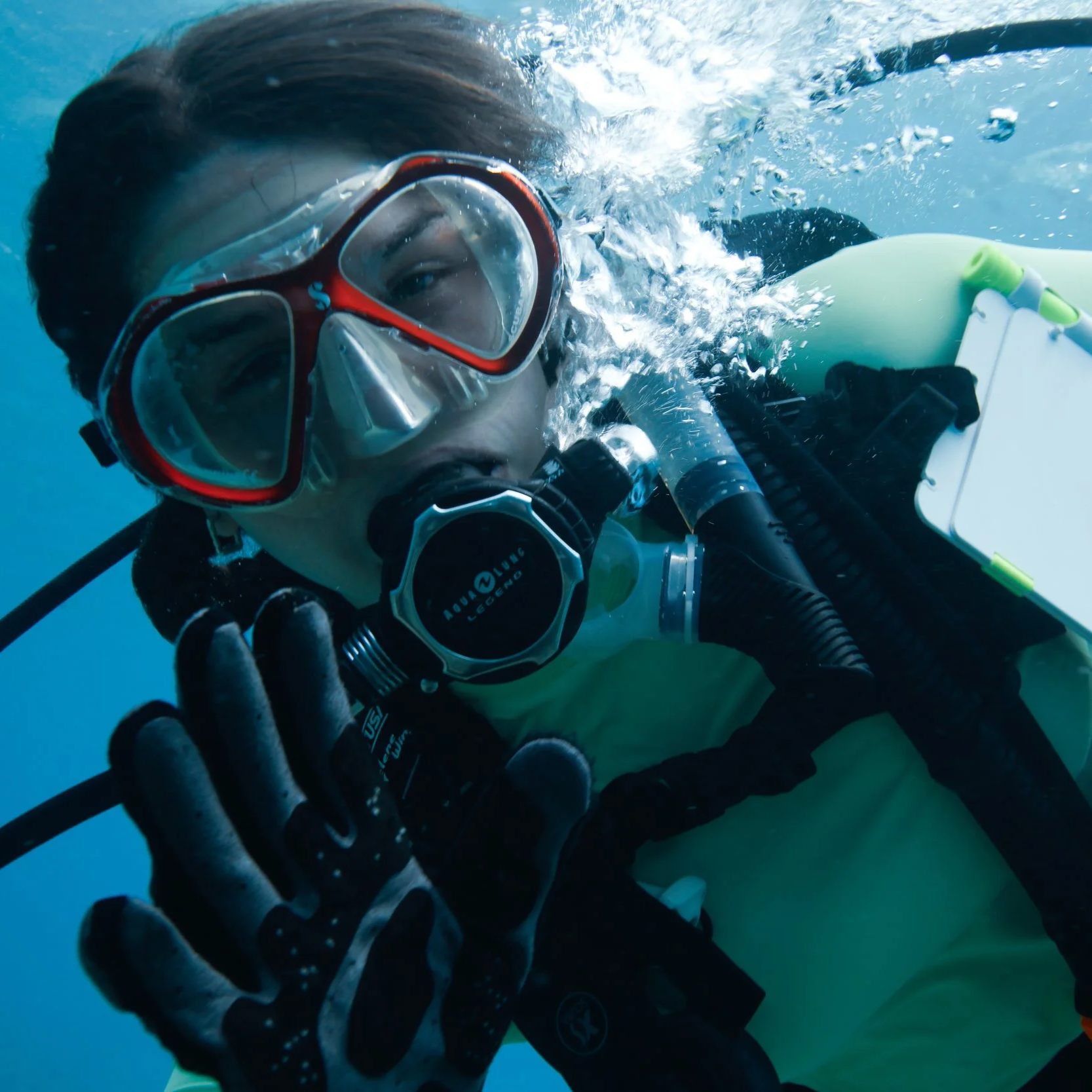An underwater photograph of a scuba diver wearing goggles and a wetsuit, holding one hand up in a stop gesture.