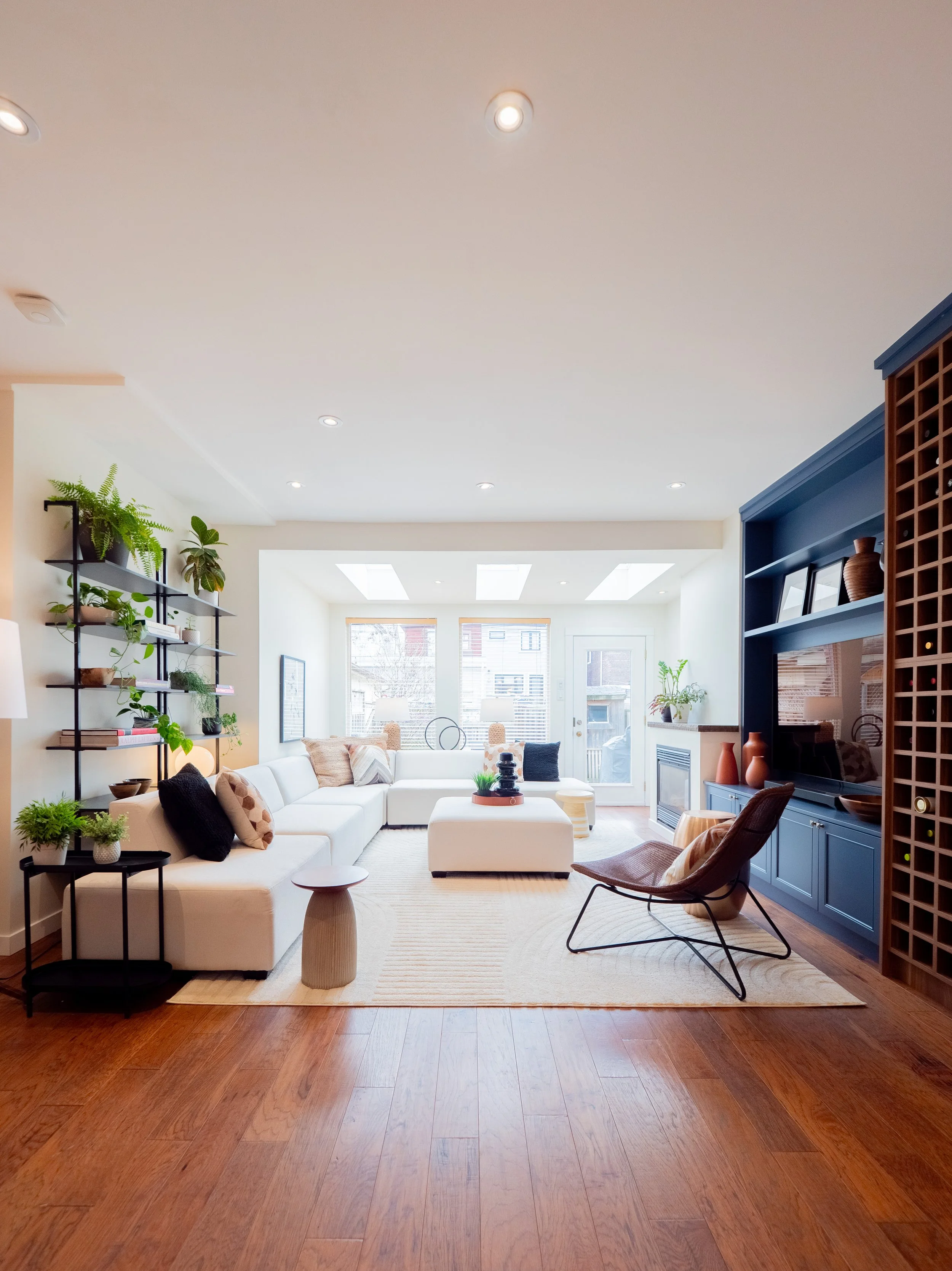 Bright living room with white sectional sofa, black and beige pillows, wooden floor, blue wall unit with decorative items, black chair with beige cushion, wall shelves with plants, large windows with sunlight.
