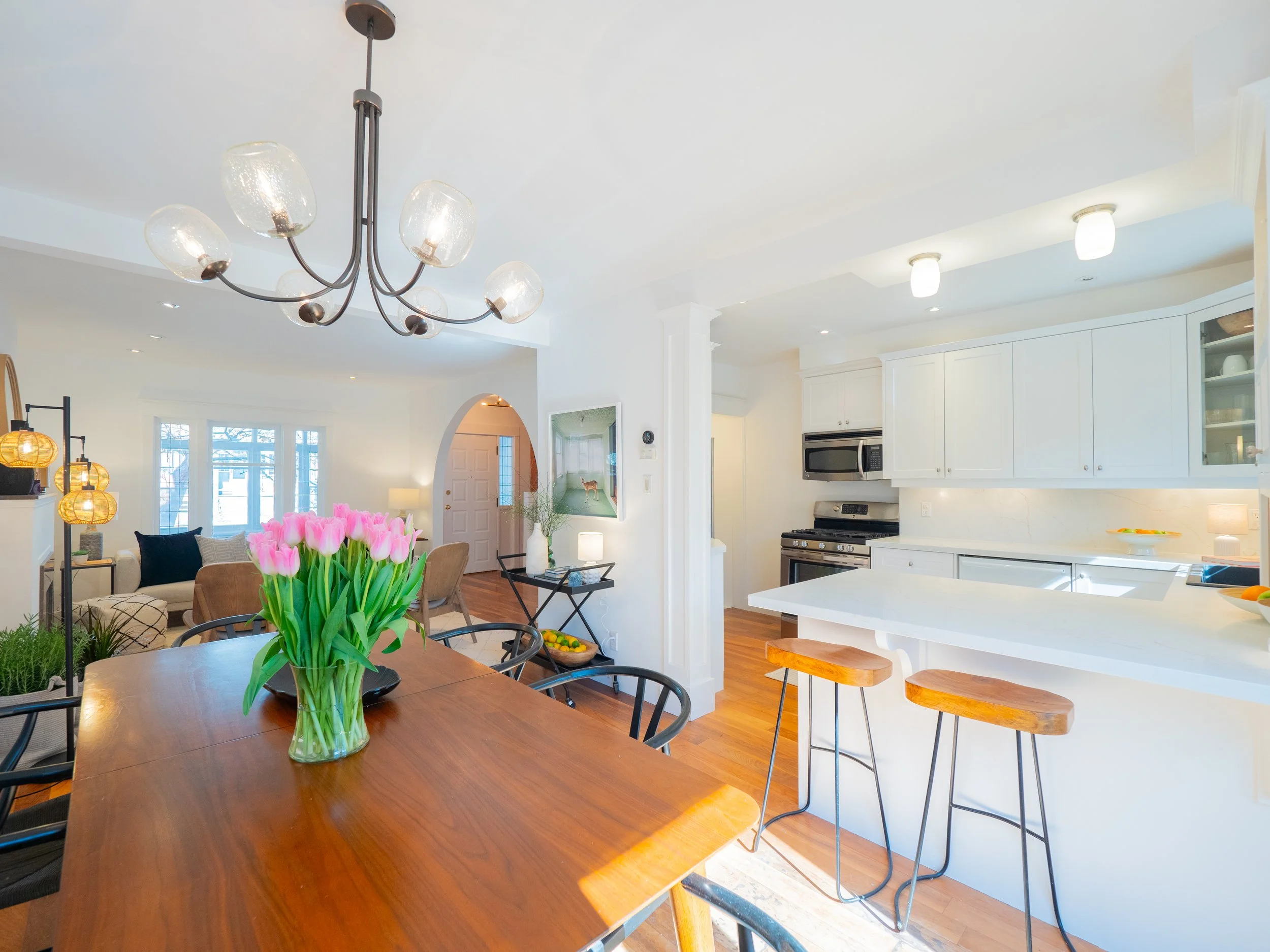Open-concept dining and kitchen area with a wooden dining table, pink tulips in a glass vase, white cabinets, and barstools at a kitchen island.