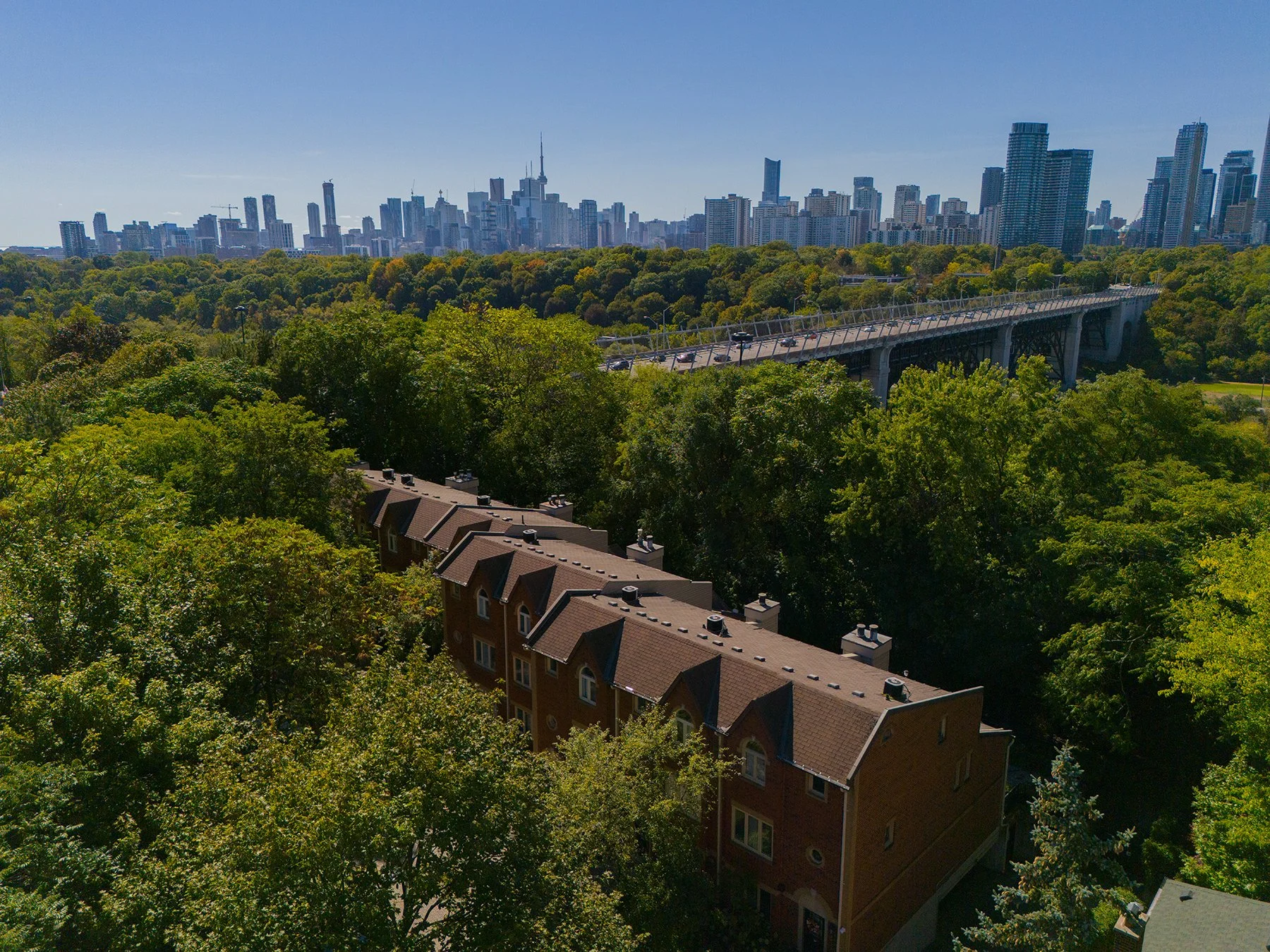 Aerial view of Toronto skyline with tall buildings in the background, a green park with dense trees in the foreground, and a bridge over the trees connecting different parts of the city from the East End Riverdale
