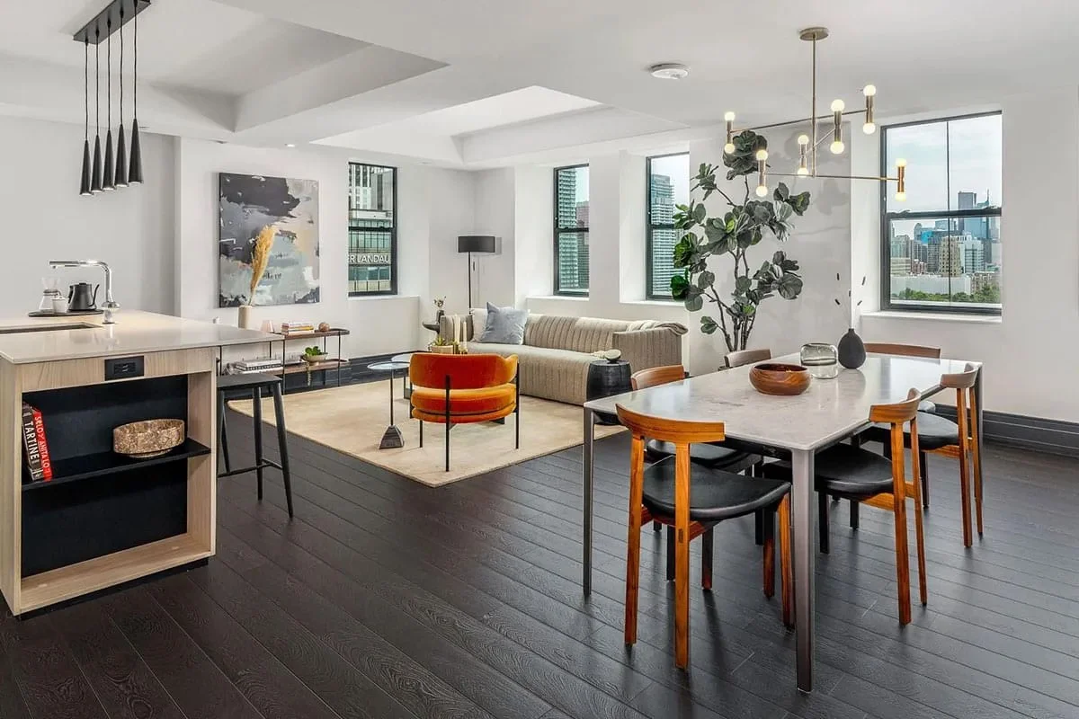 Modern open-concept living room and dining area with large windows, dark wooden flooring, a sofa, orange accent chair, dining table with eight chairs, and greenery.