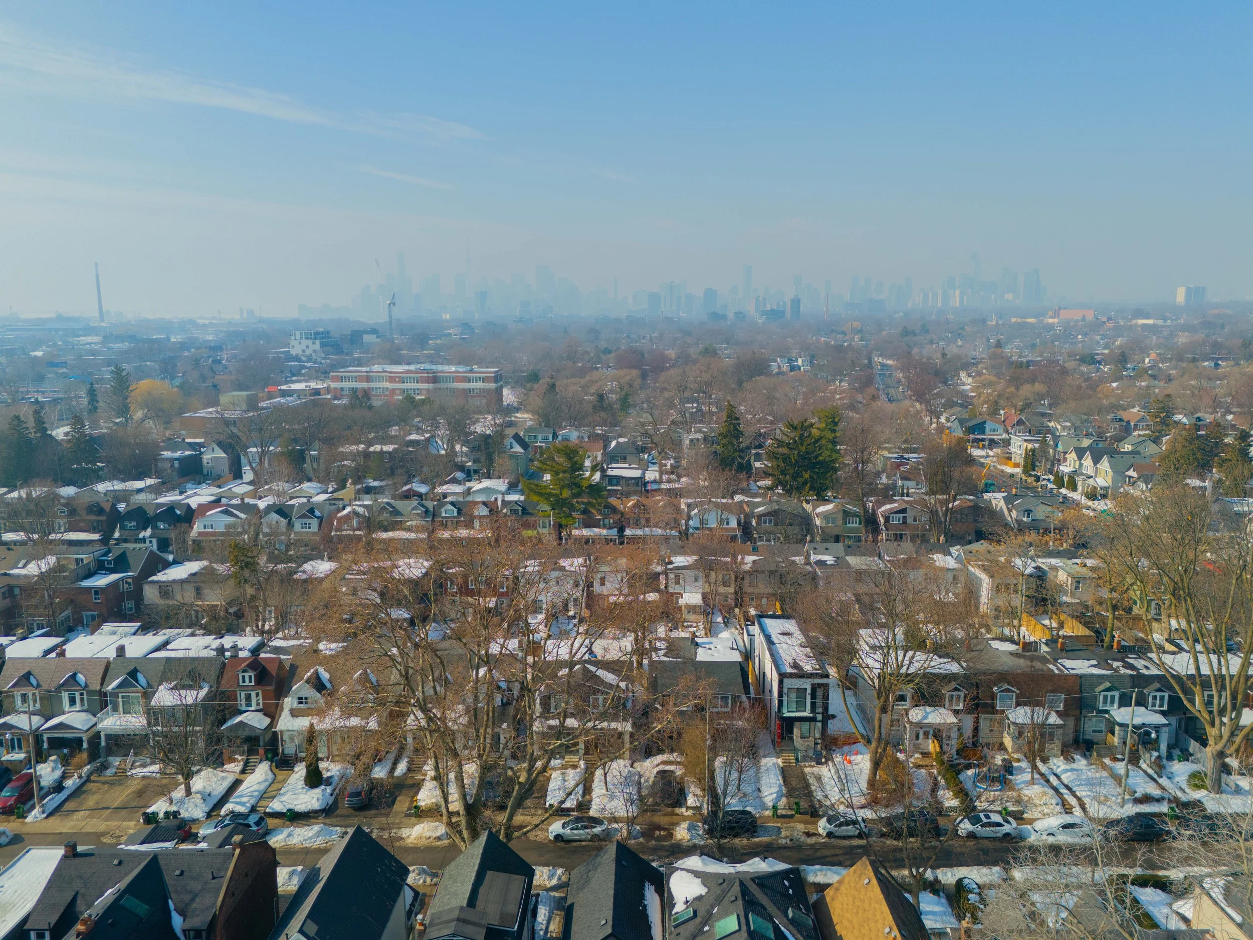 Aerial view of a residential neighborhood with snow on roofs and trees, with a city skyline in the background under a clear blue sky.